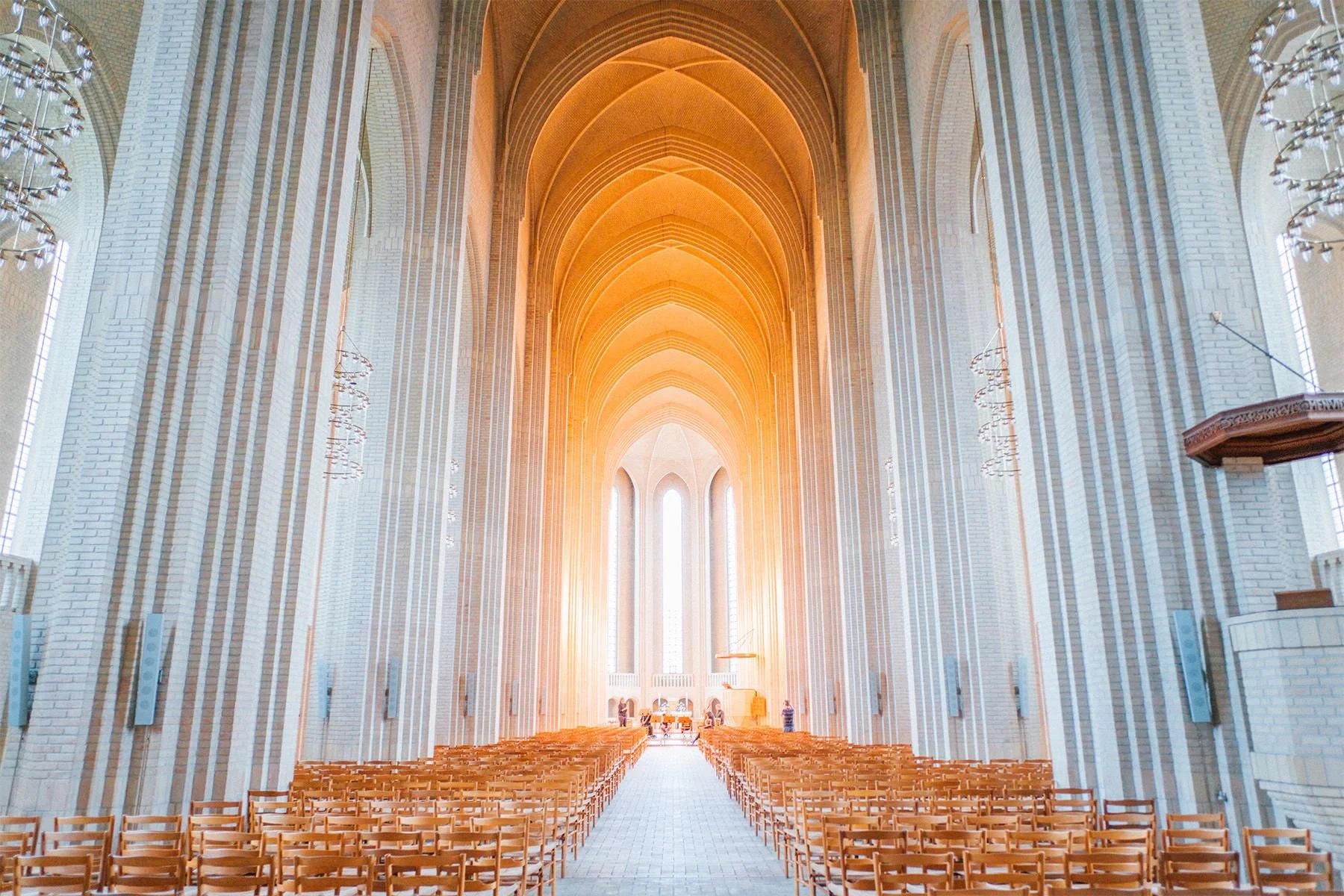Interior of a grand church or cathedral with high vaulted ceilings, tall windows, and rows of wooden chairs facing the altar at the front, illuminated by warm sunlight.