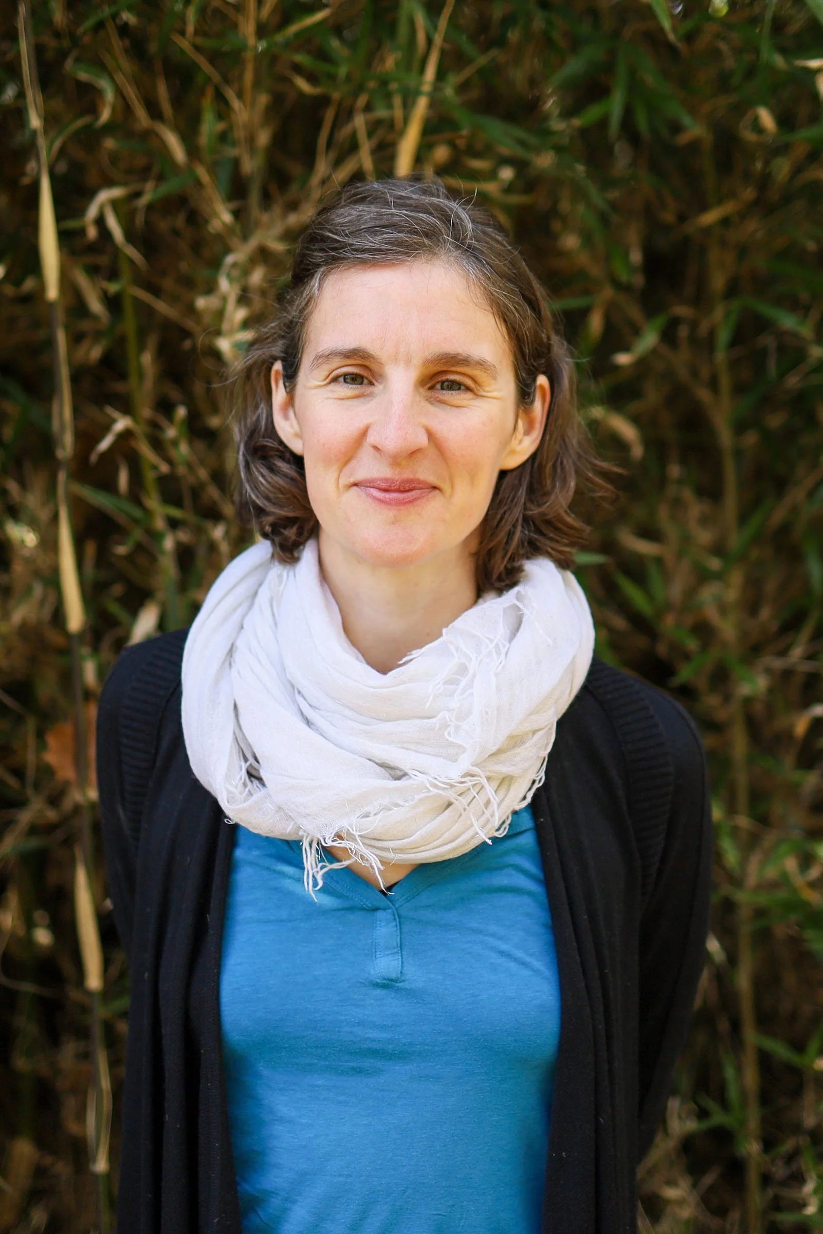 A woman with short brown hair wearing a white scarf, blue shirt, and black jacket standing outdoors in front of tall green plants.
