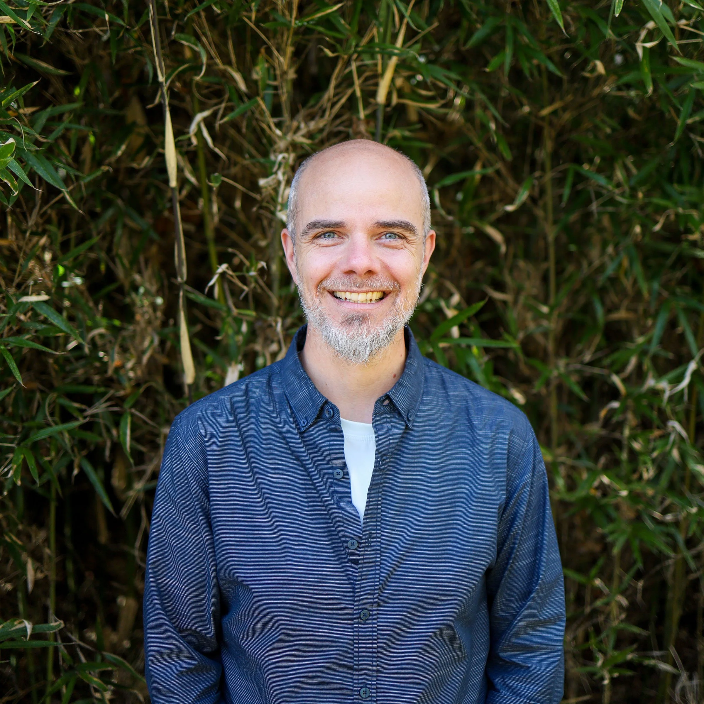 A smiling man with a beard and short hair wearing a blue button-up shirt, standing outdoors in front of green foliage.