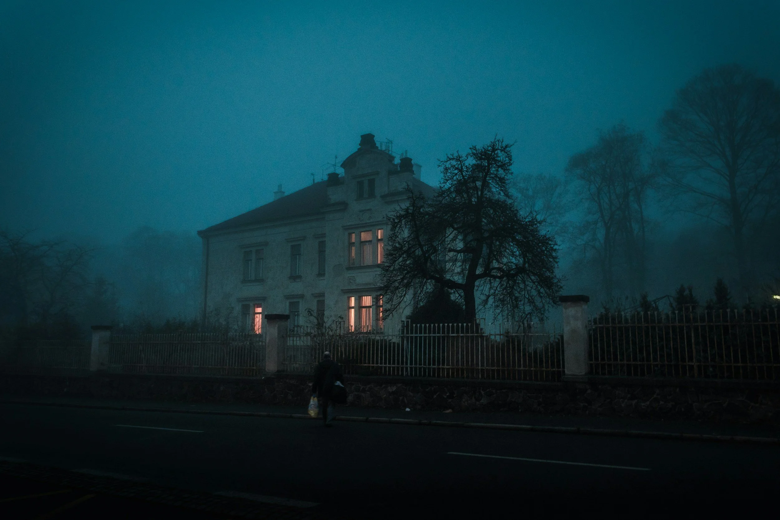 An old, eerie house with illuminated windows under a dark, foggy sky at dusk, inside which sinister happenings are hidden.