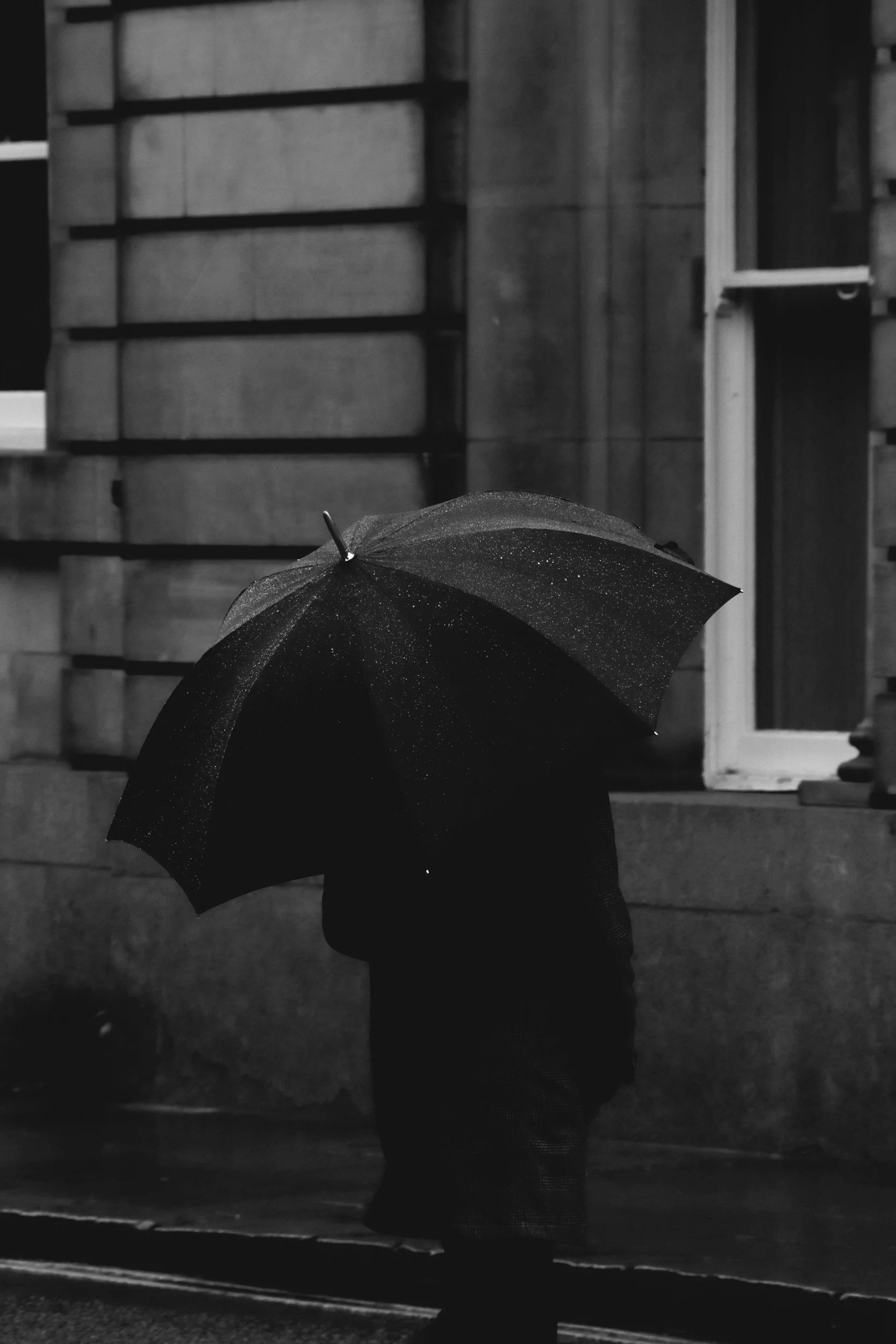 A dark figure hidden beneath a broad umbrella in the rain.