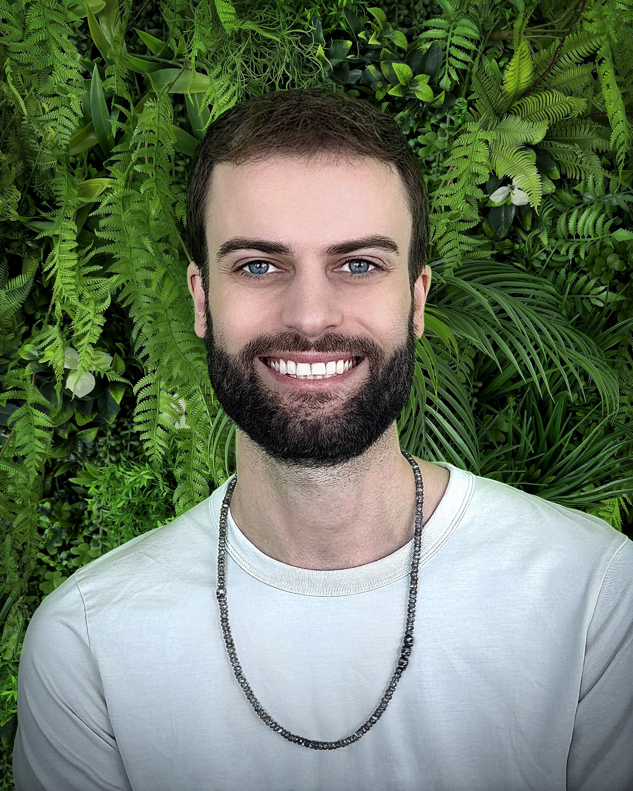 A man with blue eyes standing against a lush jungle plant wall
