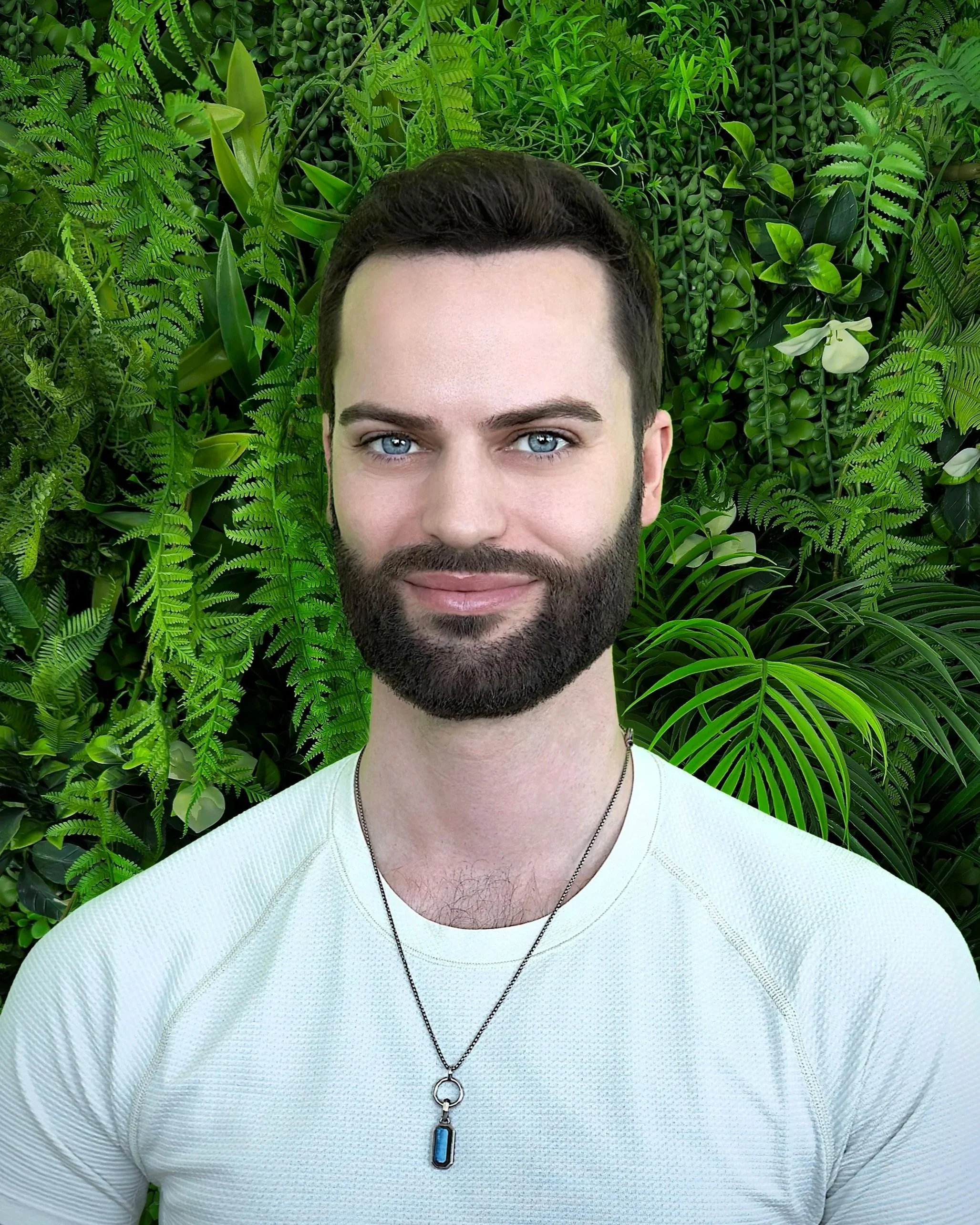A man with a beard and blue eyes smiling in front of a lush green background of various houseplants and ferns.