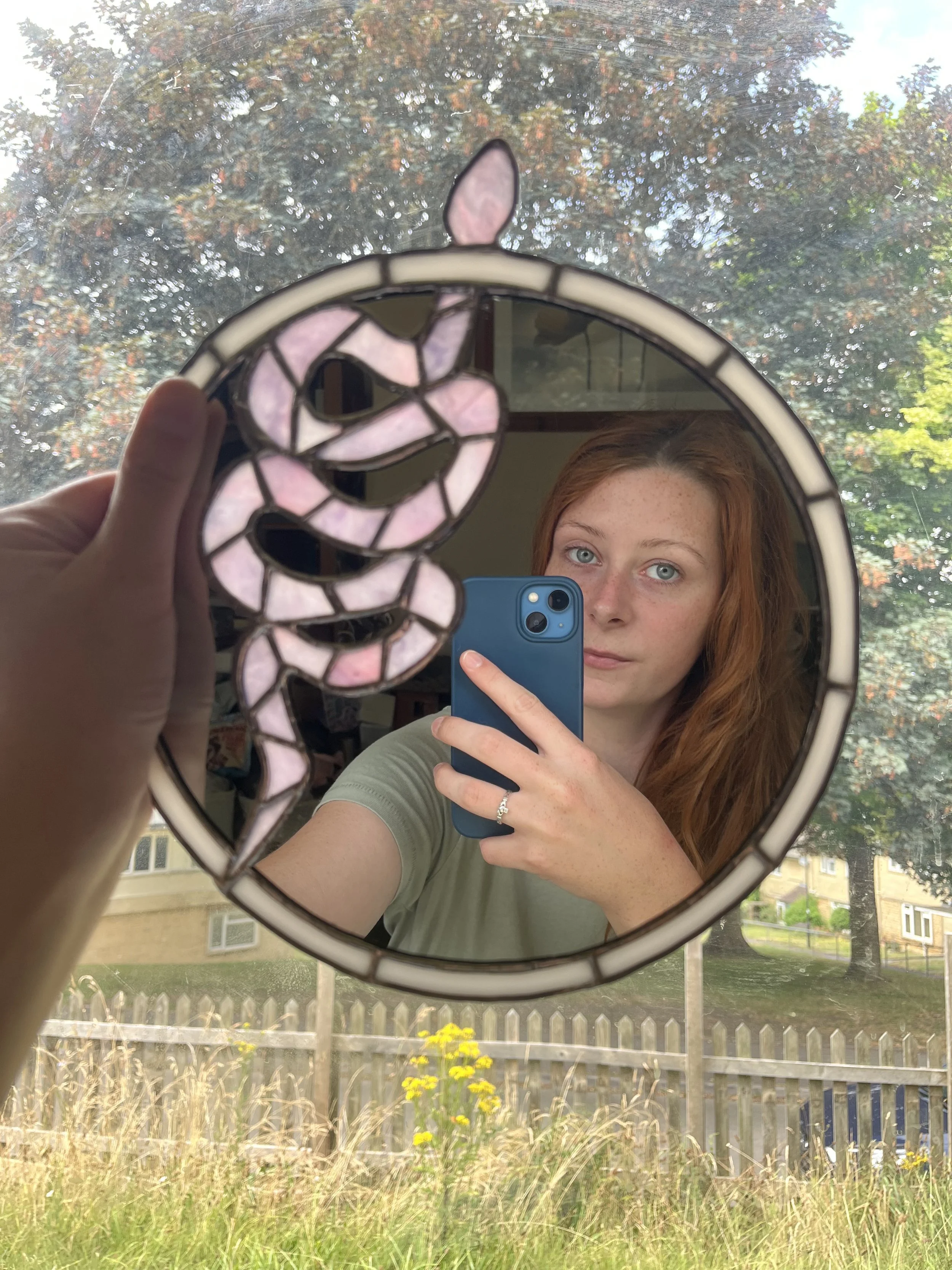 A young woman with red hair and blue eyes taking a mirror selfie with her phone, holding a stained glass snake mirror in front of her face. Outside the window, trees and a grassy garden are visible.