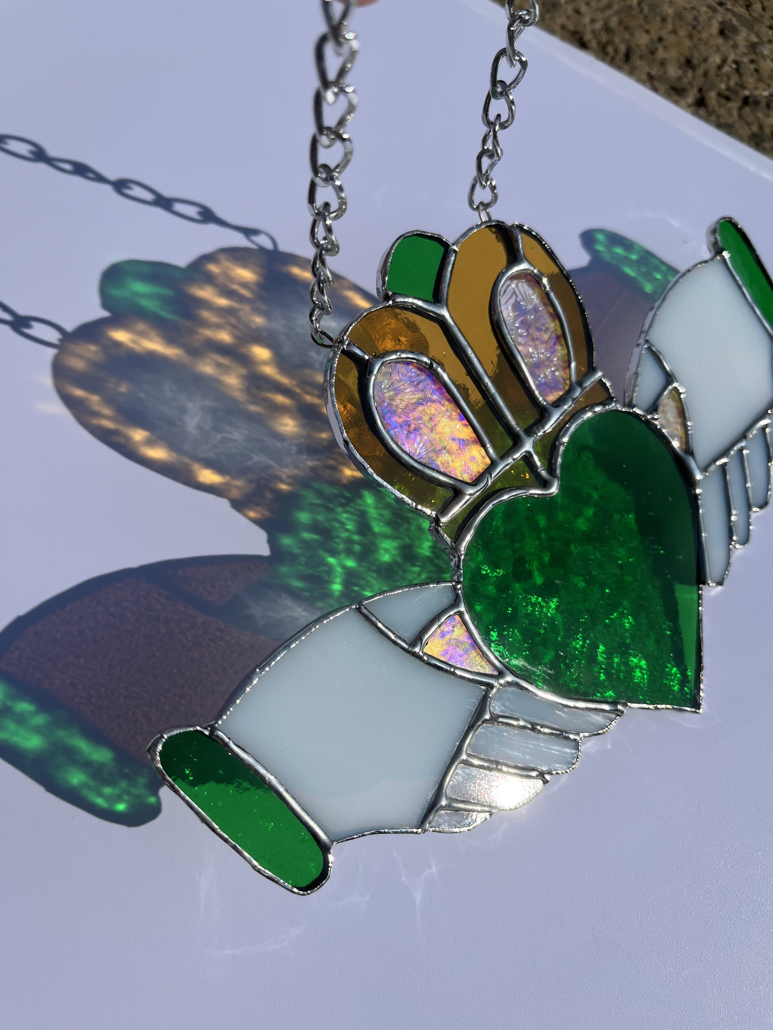 Stained glass traditional Claddagh with a green heart, pale hands, and an amber crown on top, casting colourful shadows on a white surface.