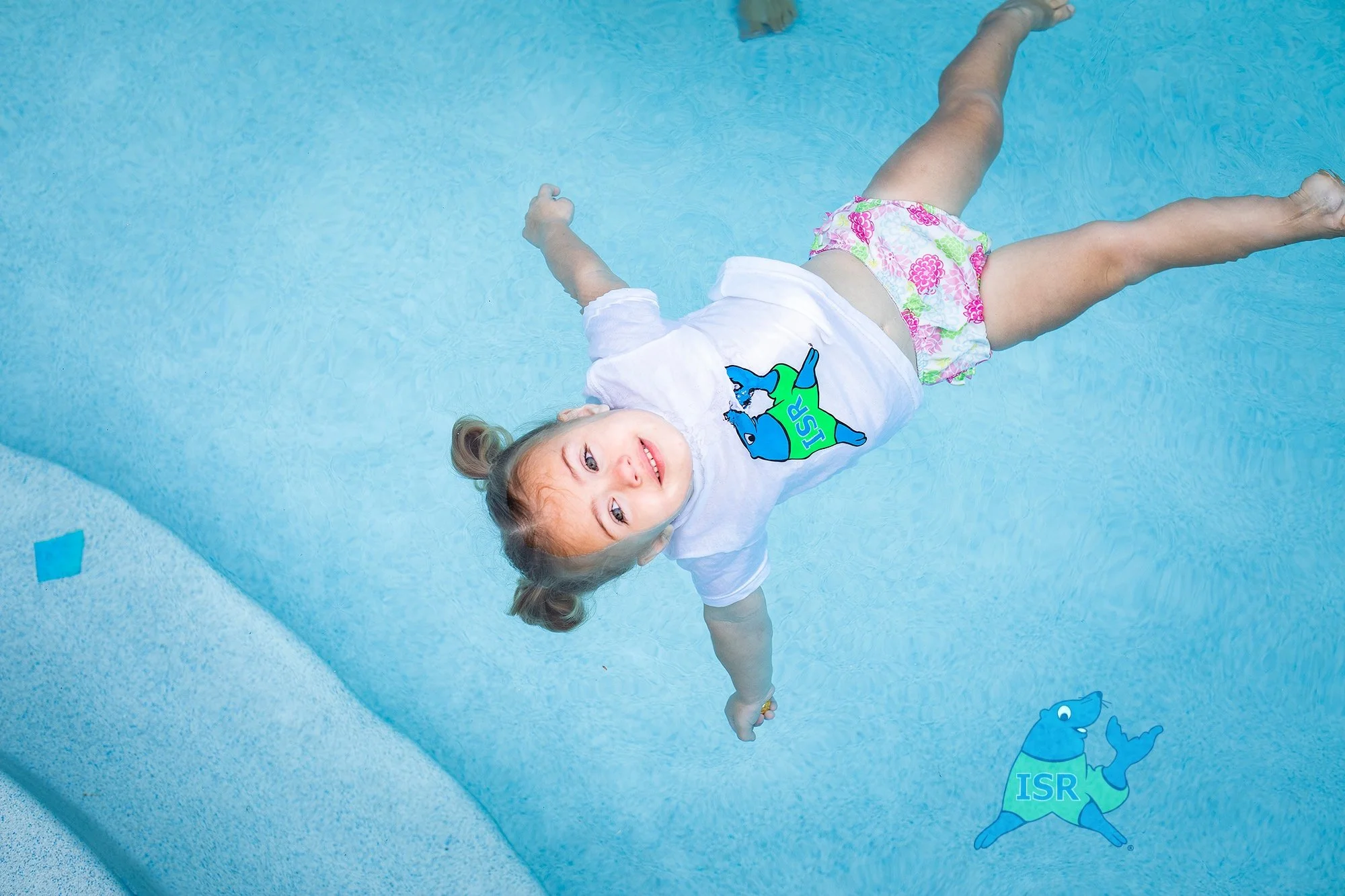 A young girl floating in a swimming pool on her back, smiling and looking at the camera, wearing a white t-shirt with a blue and green cartoon animal and pink floral swim bottoms.