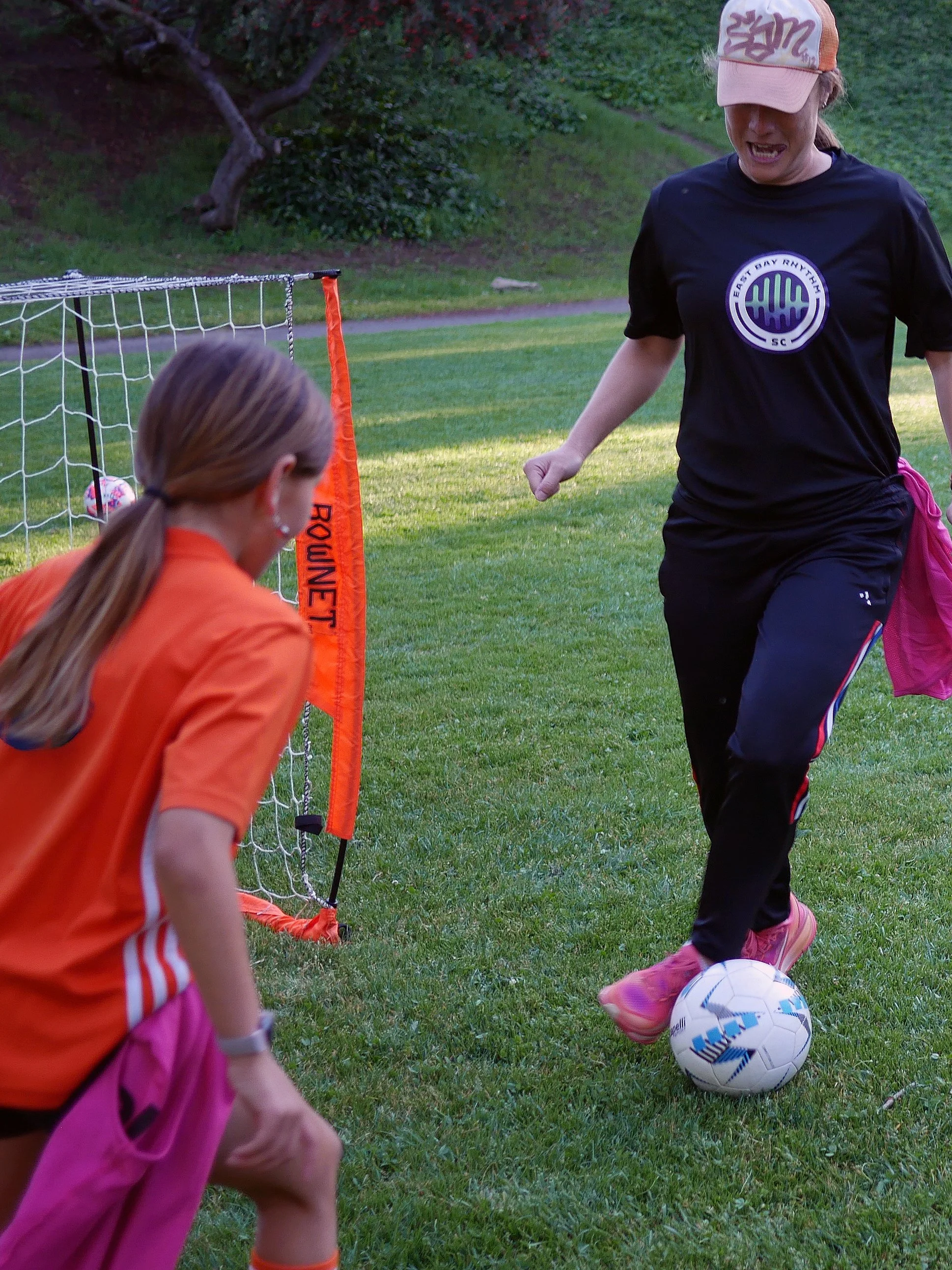 Two young girls playing soccer outdoors on a grassy field. One girl in an orange jersey and pink shorts is watching while the other girl, wearing a black shirt and pink shoes, is kicking a soccer ball near a small goal.