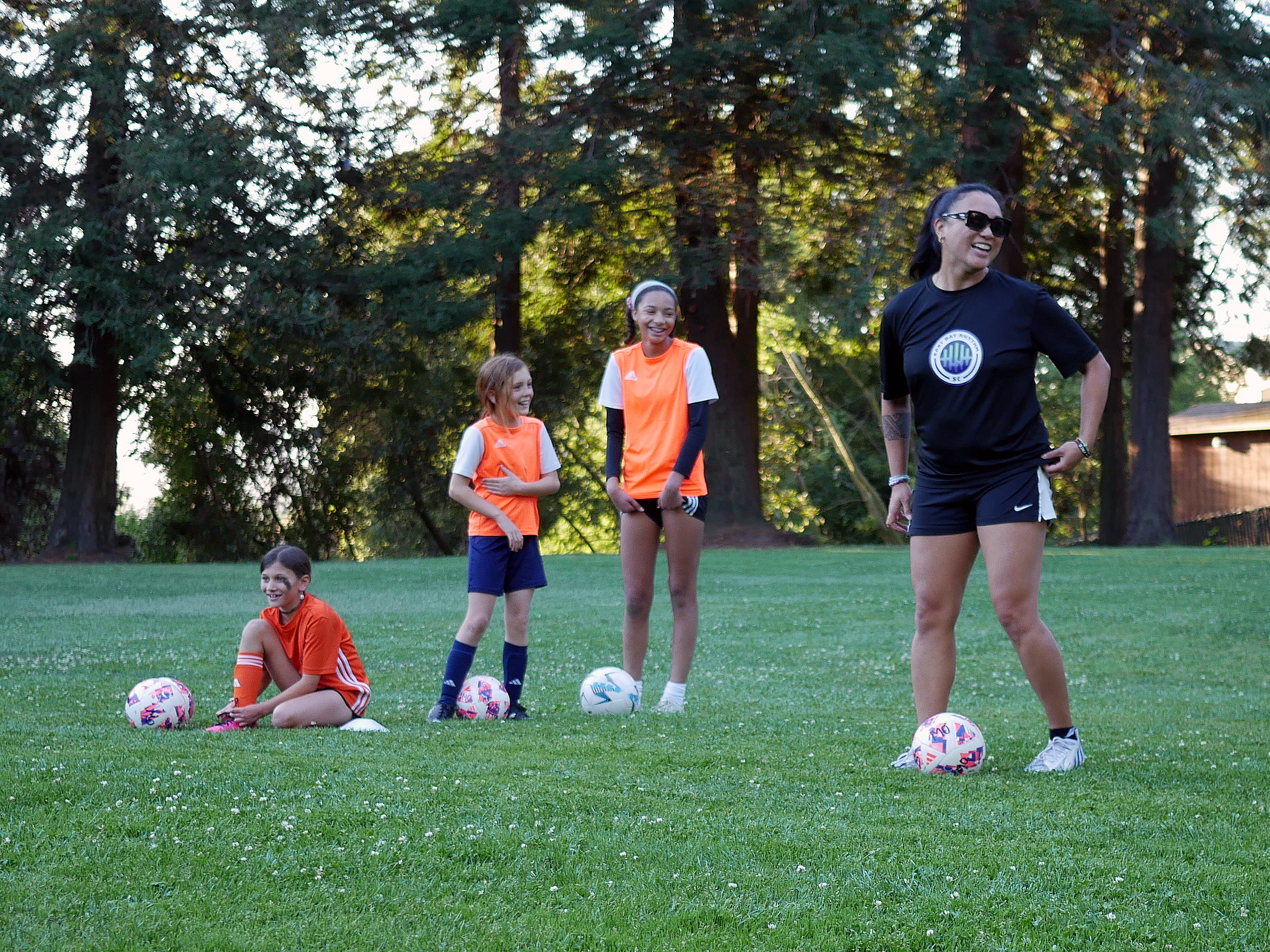 A woman and three young girls on a soccer field, smiling and standing with soccer balls in front of tall trees, during the daytime.