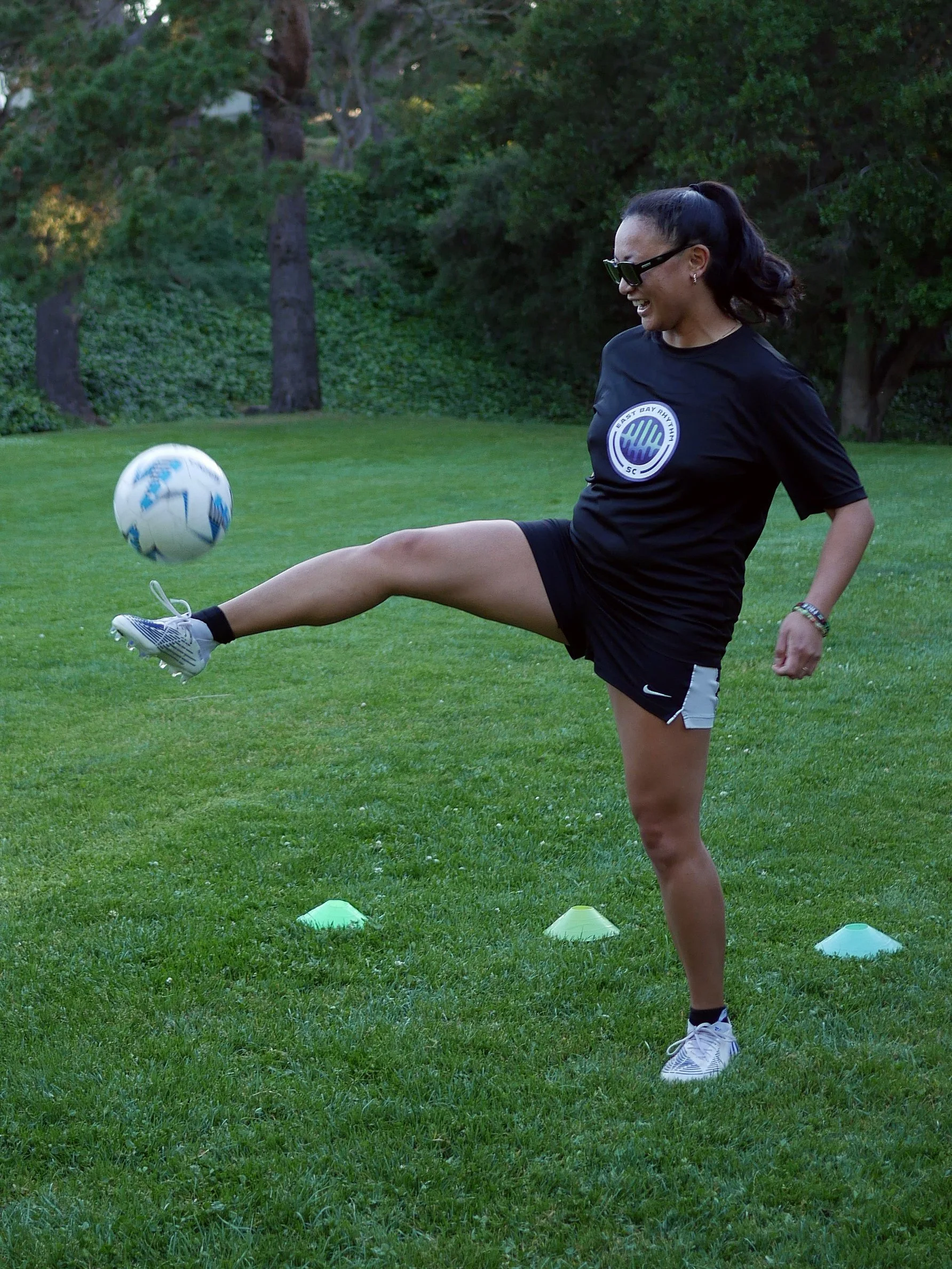 A woman playing soccer outdoors on a grassy field, kicking a soccer ball, surrounded by trees and colorful cones.