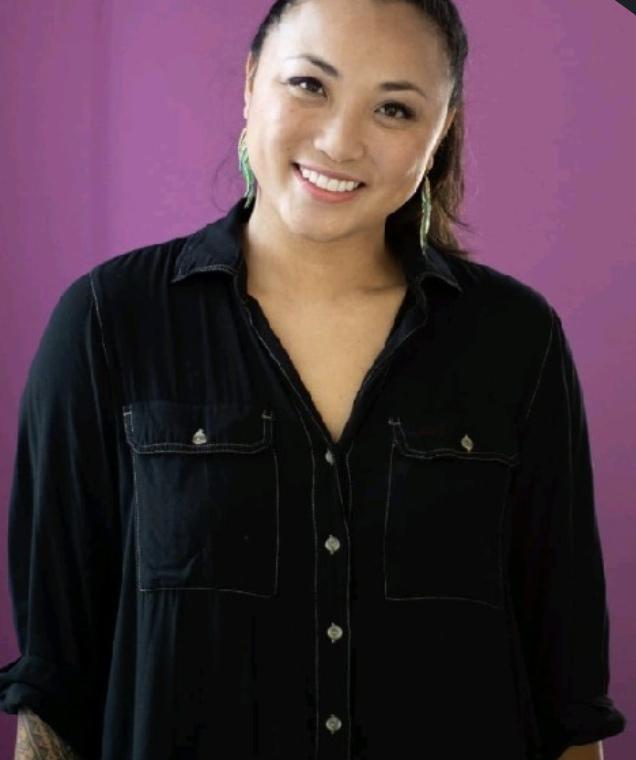 A woman smiling, wearing a black shirt with rolled-up sleeves and green earrings, standing against a purple background.