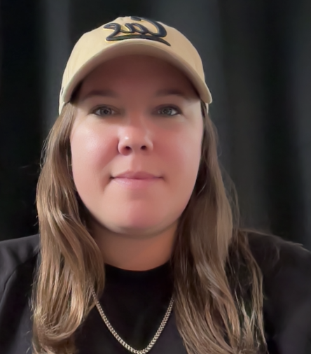A young woman wearing a yellow baseball cap and a black shirt, with a gold chain necklace, smiling at the camera against a dark background.