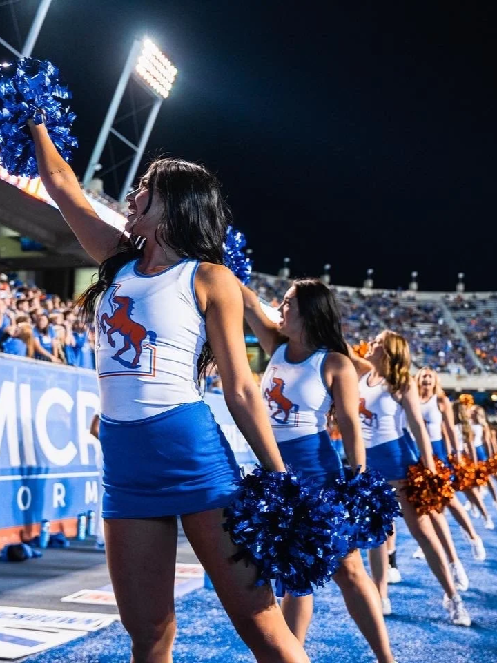 A group of cheerleaders in blue and white uniforms with a horse logo, holding blue and orange pom-poms, cheer at a sports event in a stadium at night.