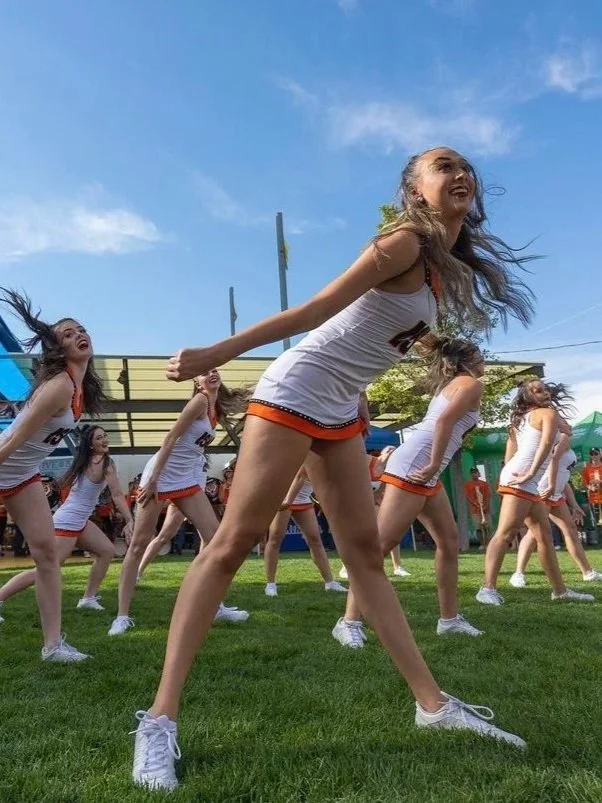 A group of cheerleaders in white and orange uniforms performing a dance on a grassy field outdoors on a sunny day.