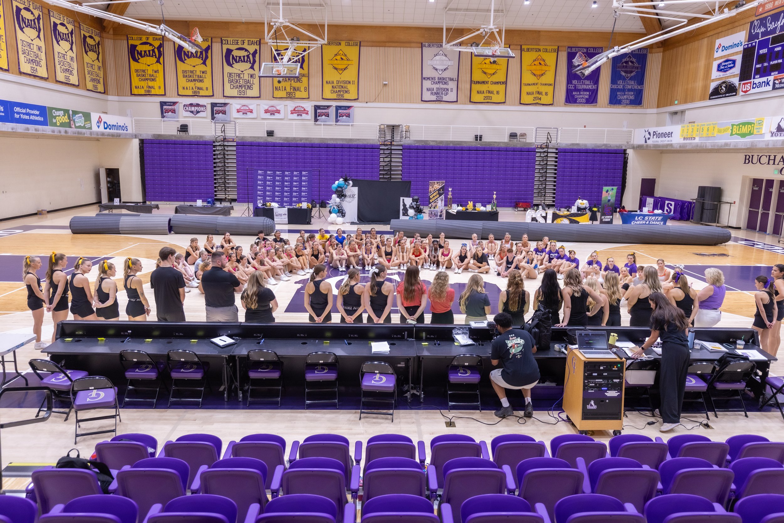 Cheerleaders and coaches gathered in a gymnasium, sitting in a circle on the floor, for a team meeting or practice. The gym has purple seats and banners hanging from the ceiling. Equipment and decorations are set up around the gym.