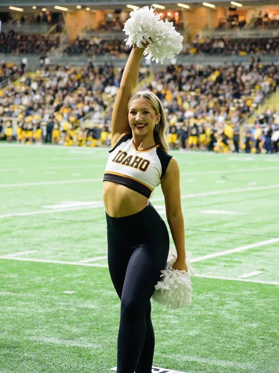 A cheerleader in a black and yellow uniform with 'IDAHO' on it, holding white pom-poms, stands on a football field in a stadium filled with spectators.