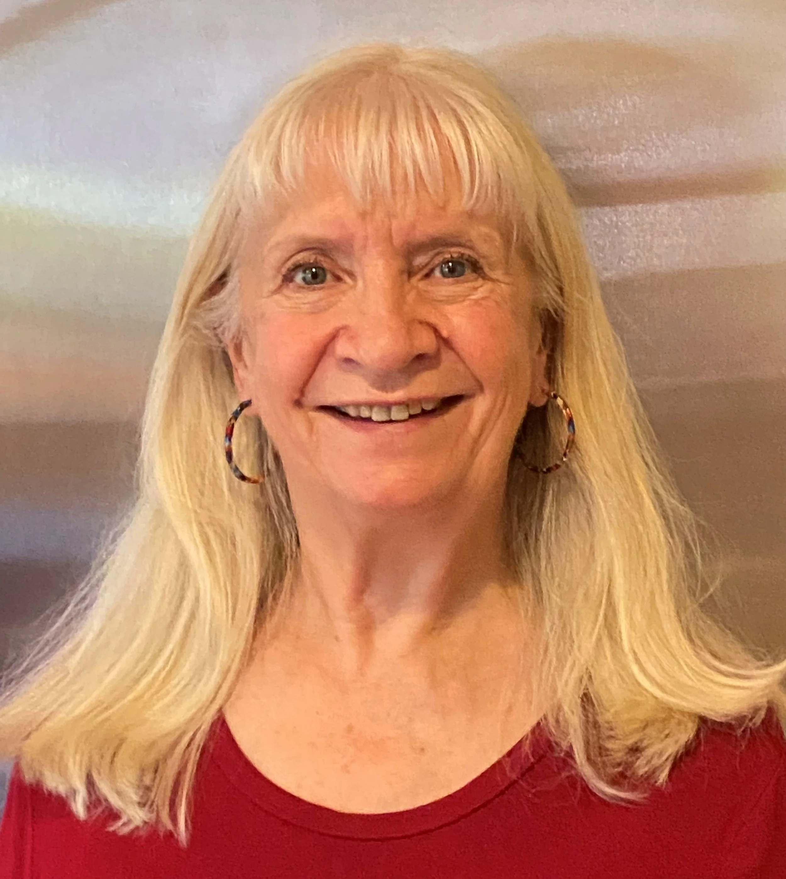 Portrait of a middle-aged woman with blonde hair, blue eyes, wearing hoop earrings and a red top, smiling in front of a colorful, blurred background.