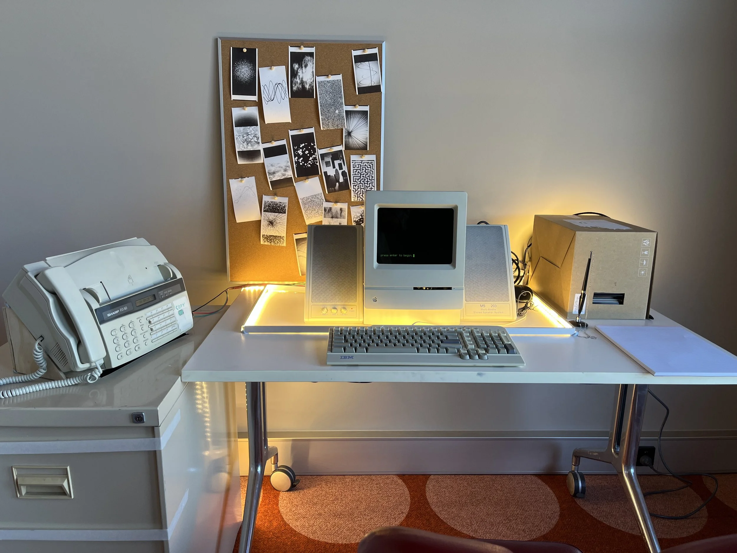 An old macintosh computer on a desk, on the desk is some strip lighting, an old keyboard, a pen and paper, a cardboard box and a pin board. An old fax machine sits on a filing cabinet