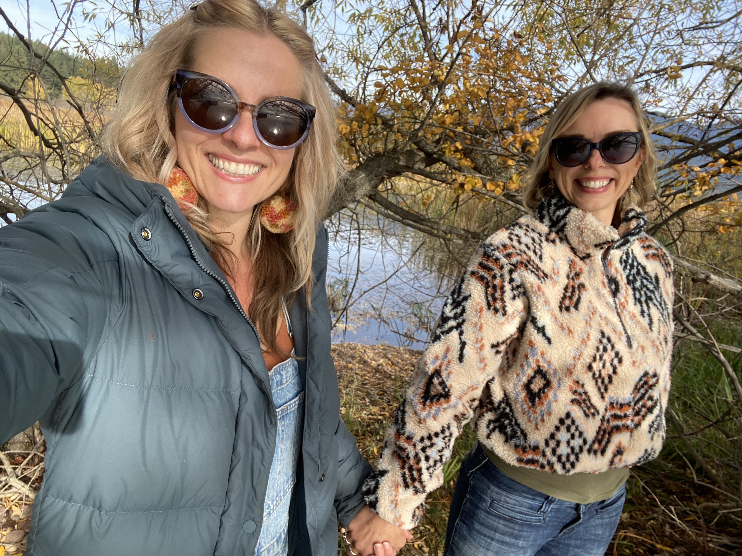 Two women smiling and holding hands outdoors near a river with trees showing fall foliage in the background.