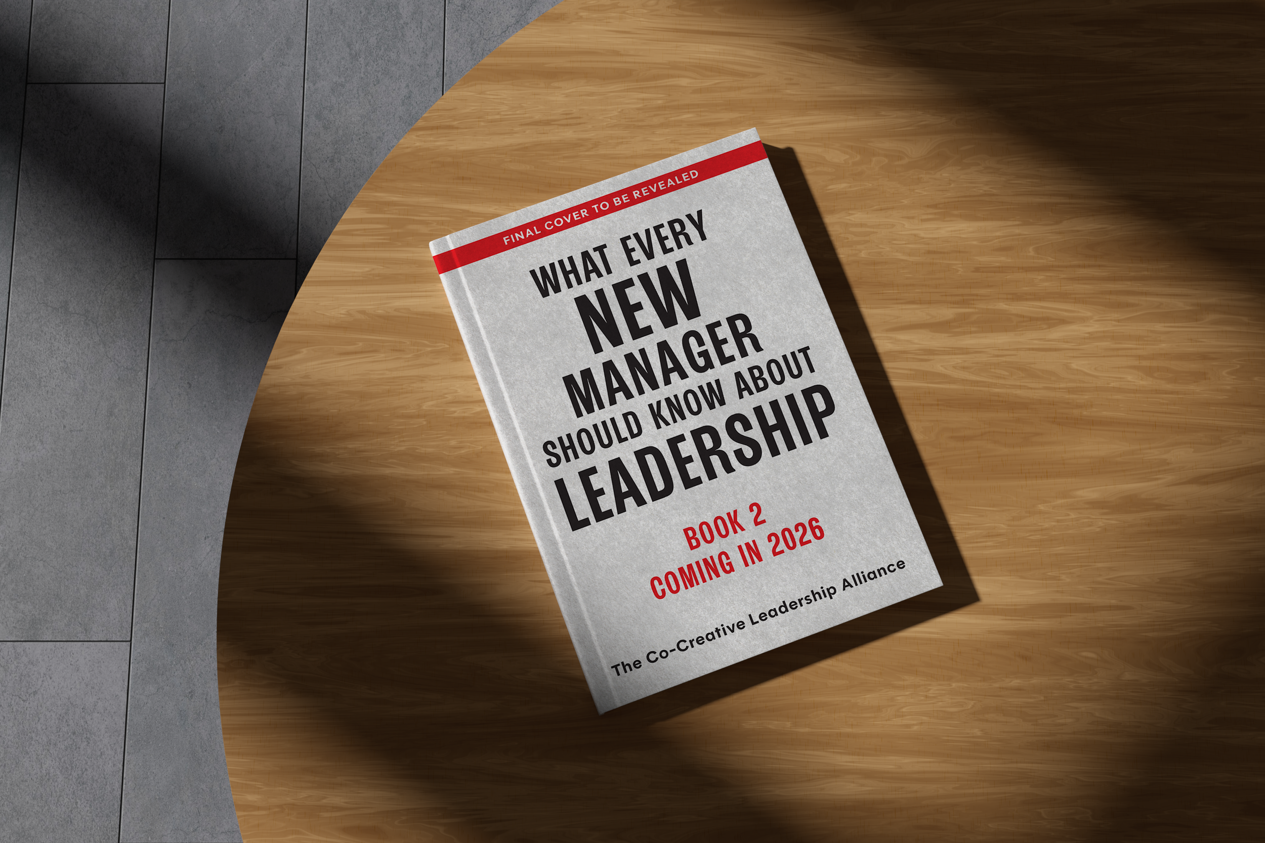 A book titled "What Every New Manager Should Know About Leadership Book 2 Coming in 2026" lies on a wooden surface, with a shadow cast over part of it, and a tiled floor nearby.