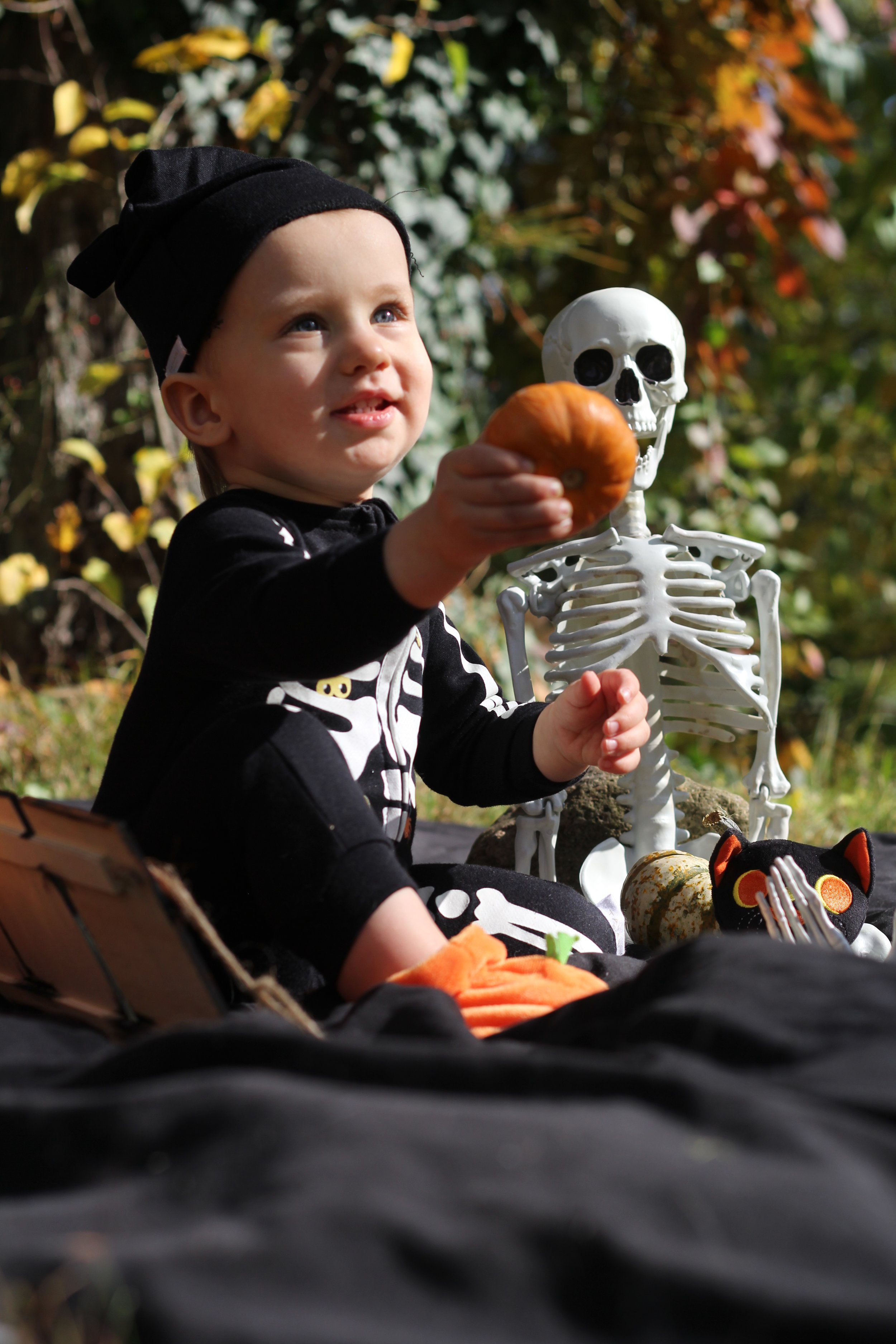 Child dressed in skeleton costume holding a pumpkin, sitting outdoors with Halloween decorations including a skeleton and black cat plush.