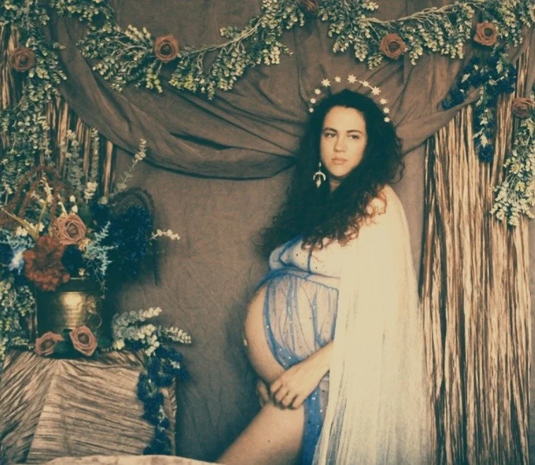 A pregnant woman with long curly hair standing in a decorated setting with flowers and drapery, posing with a serious expression.