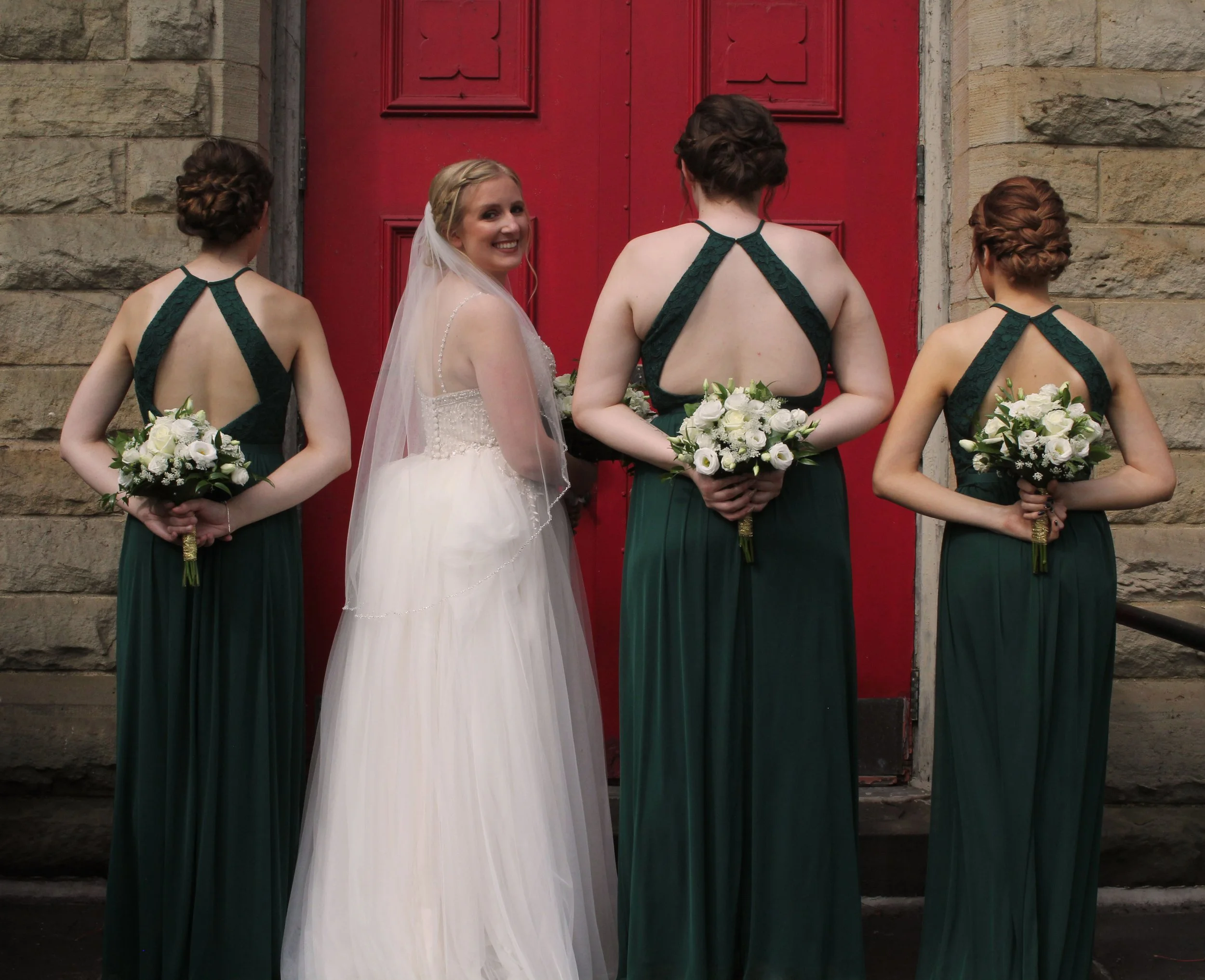 Bride in white wedding dress and veil standing with three bridesmaids in dark green dresses holding bouquets, outside in front of a red door.