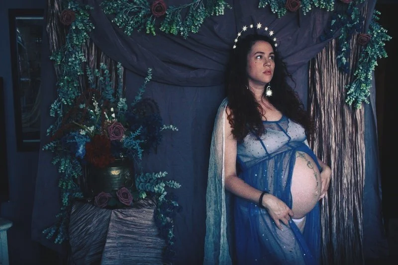 Pregnant woman with dark hair wearing a sheer blue gown and crown of stars, standing in front of a dark purple or blue curtain decorated with greenery and flowers, next to a floral arrangement in a gold vase.