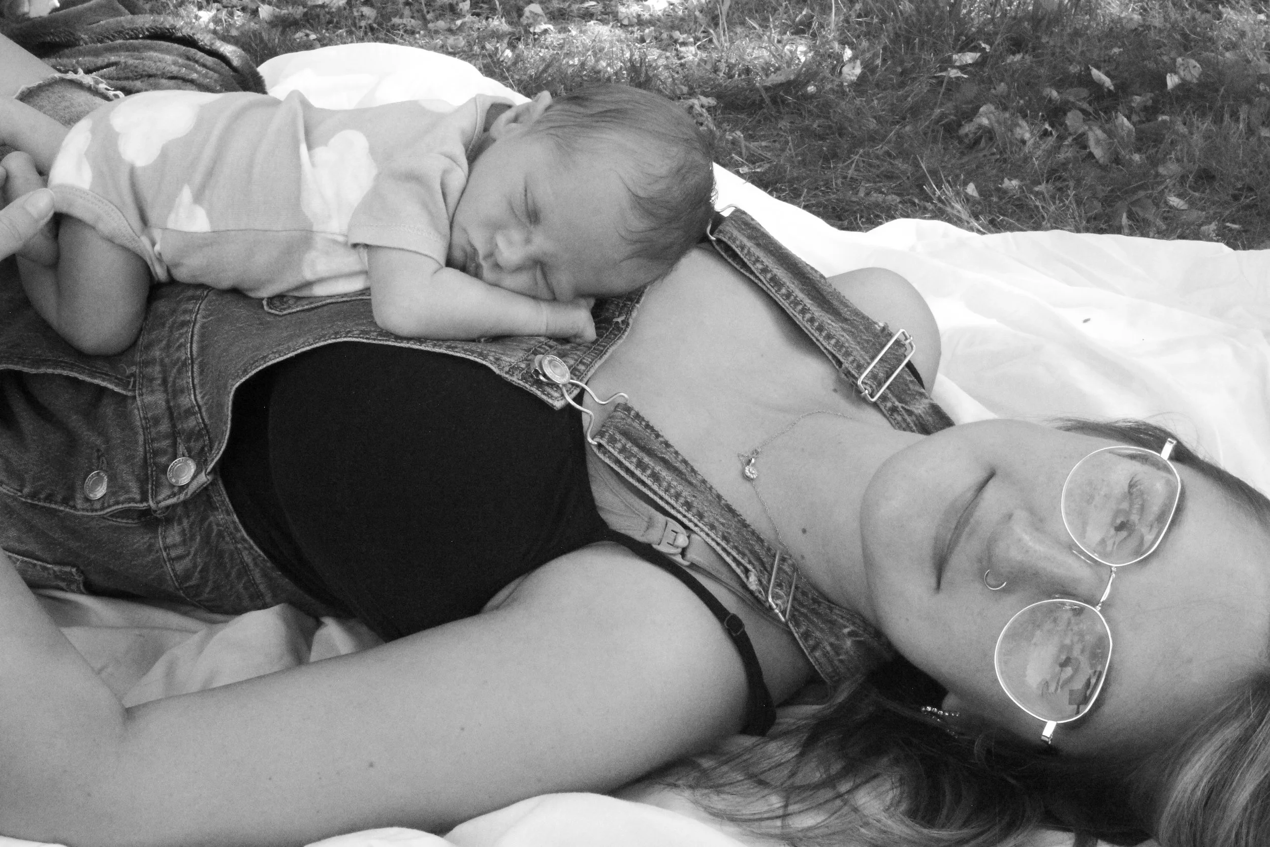 A woman lying on her back outdoors with a sleeping baby resting on her chest.