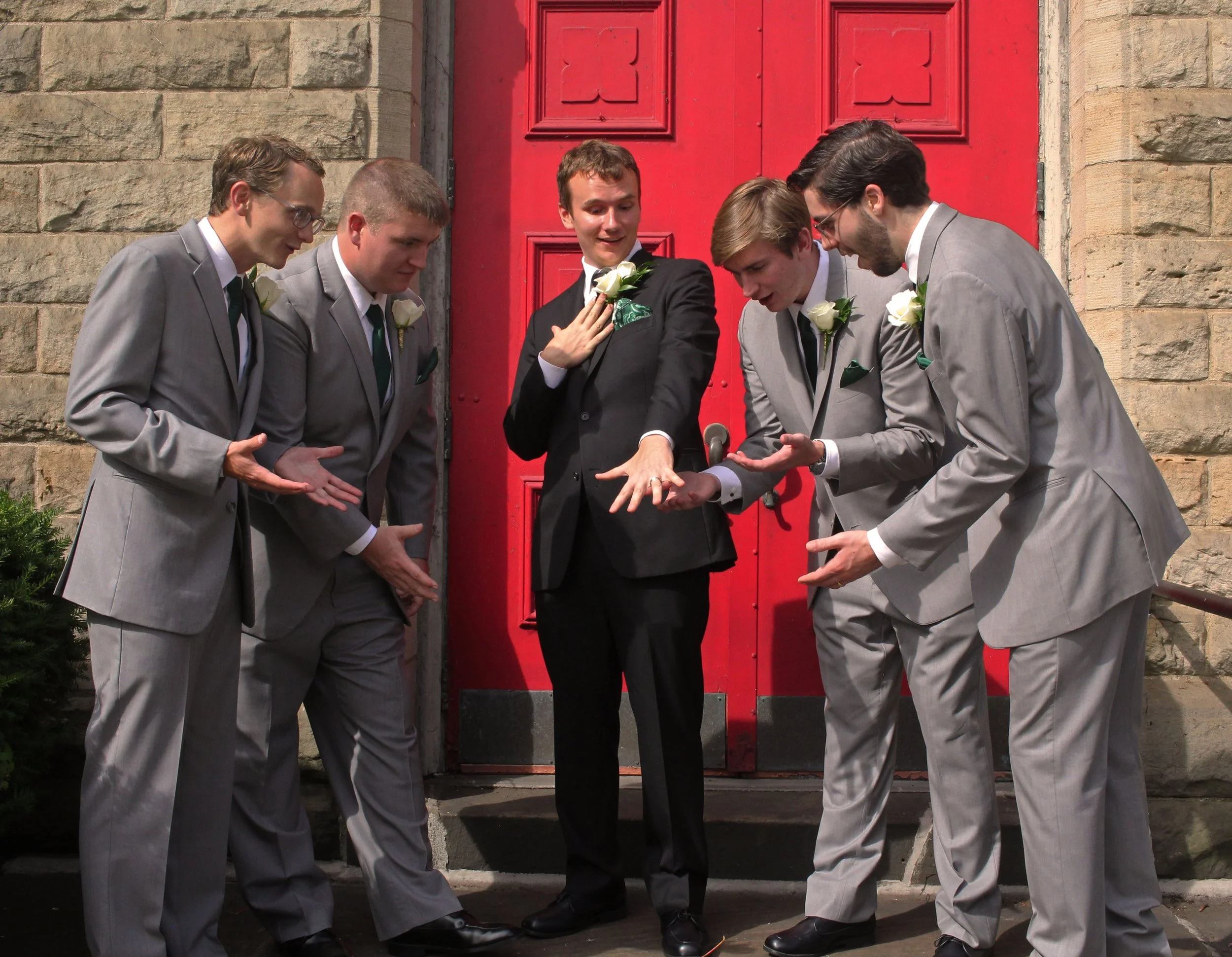 Six men in suits, five in gray and one in black, are standing outside in front of a red door with stone walls. The man in the black suit is showing a ring on his finger to the others, who are looking at it with interest.