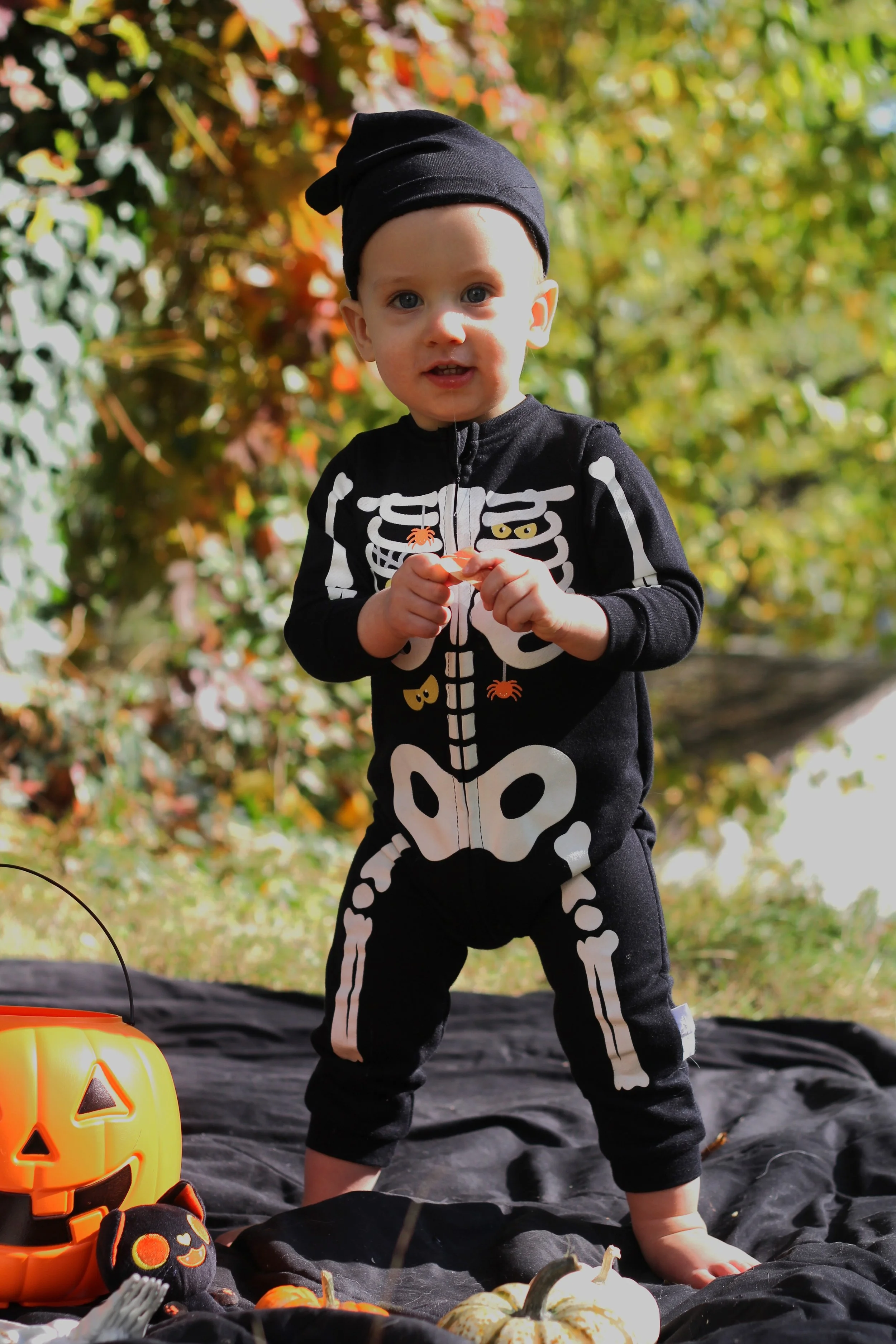 Young child dressed in a black skeleton costume holding a small pumpkin, standing outdoors with a pumpkin-shaped container nearby for trick-or-treating, surrounded by fall decorations and foliage.