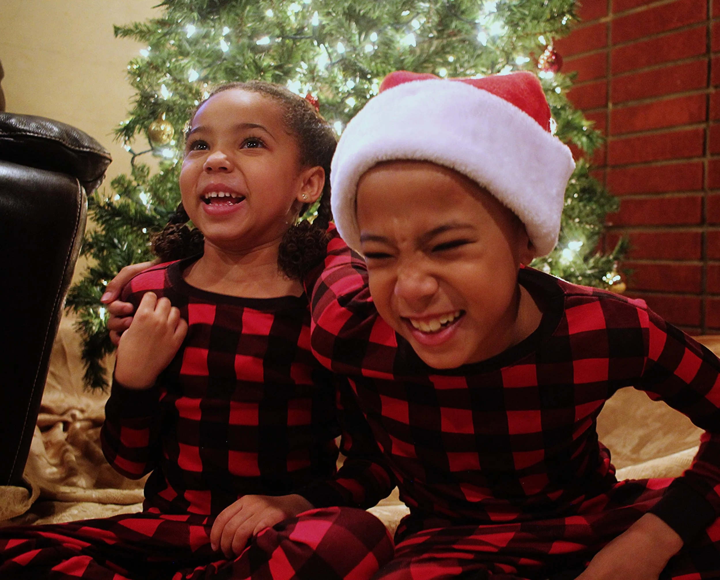 Two children in red and black plaid pajamas, celebrating Christmas in front of a decorated tree. One girl has braids and is smiling, the other boy is wearing a Santa hat and is laughing.
