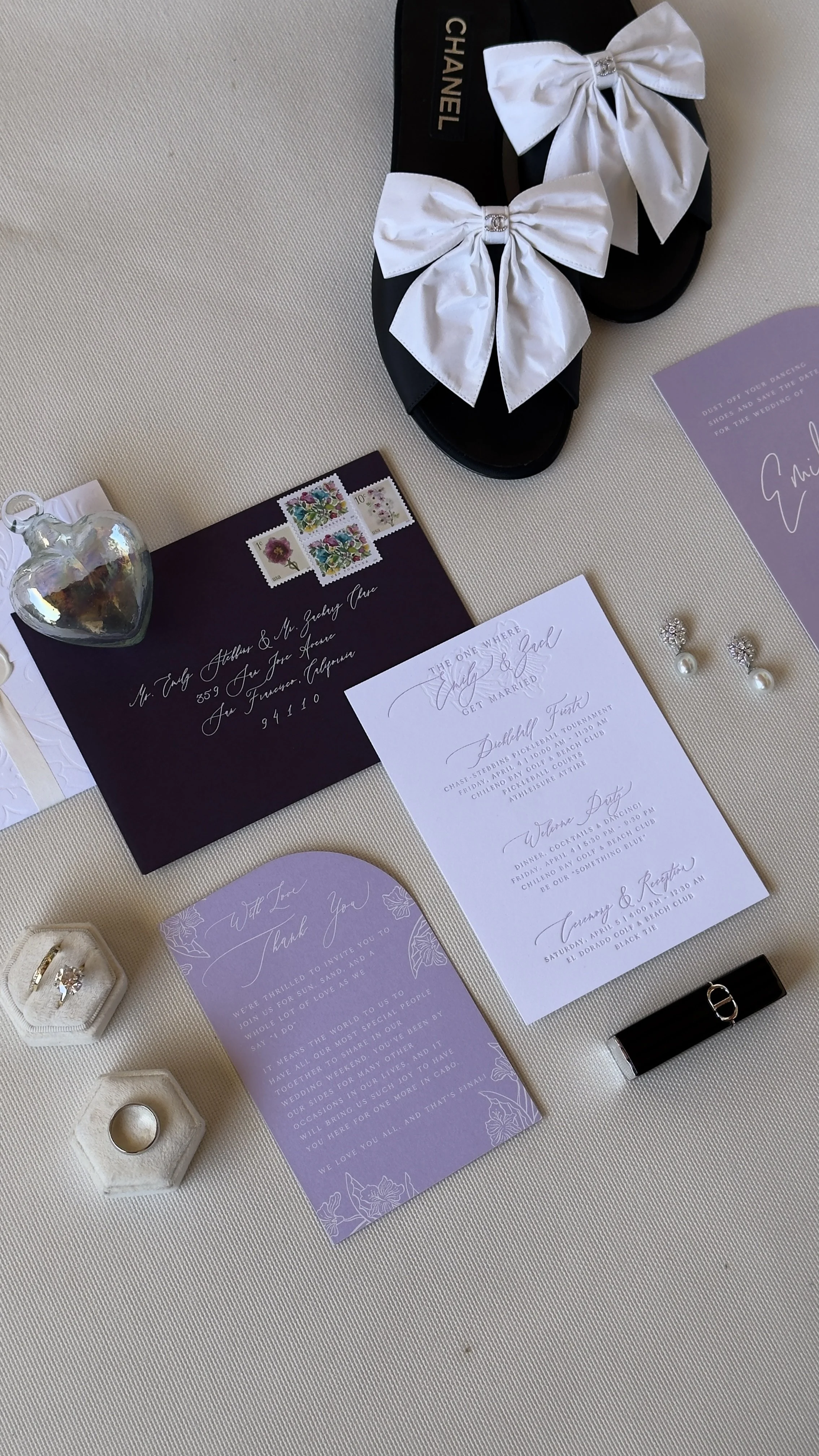 Wedding invitation suite, white satin bows on black slides, pearl earrings, wedding rings in a white velvet box, purple thank you card, purple event details card, iridescent heart-shaped ornament, black watch, and purple invitation card with wedding