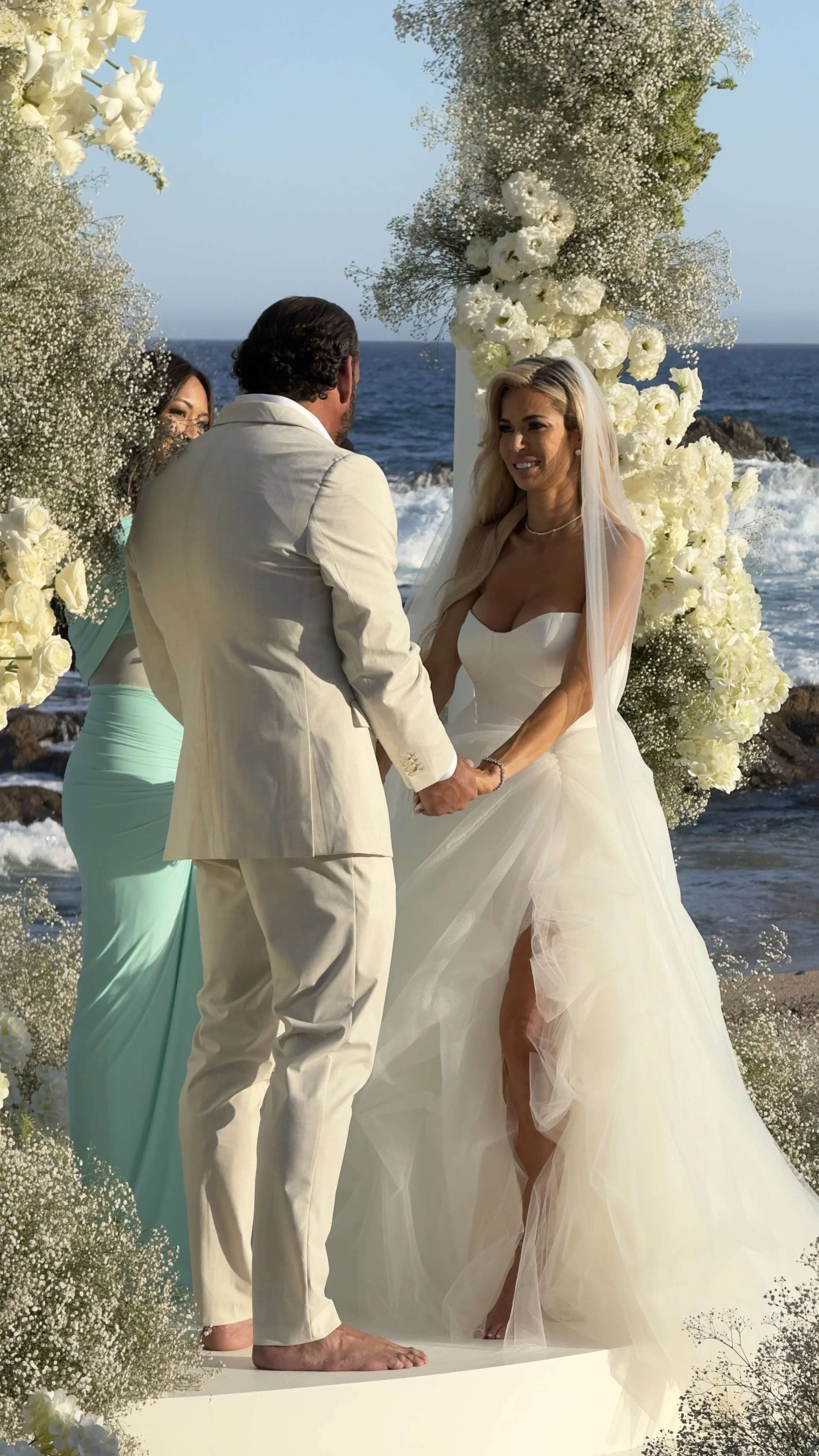 A wedding ceremony takes place on a beach, with a bride and groom holding hands and facing each other. The bride wears a strapless white gown with a slit, and the groom wears a light-colored suit. They stand beneath a floral arch decorated with white
