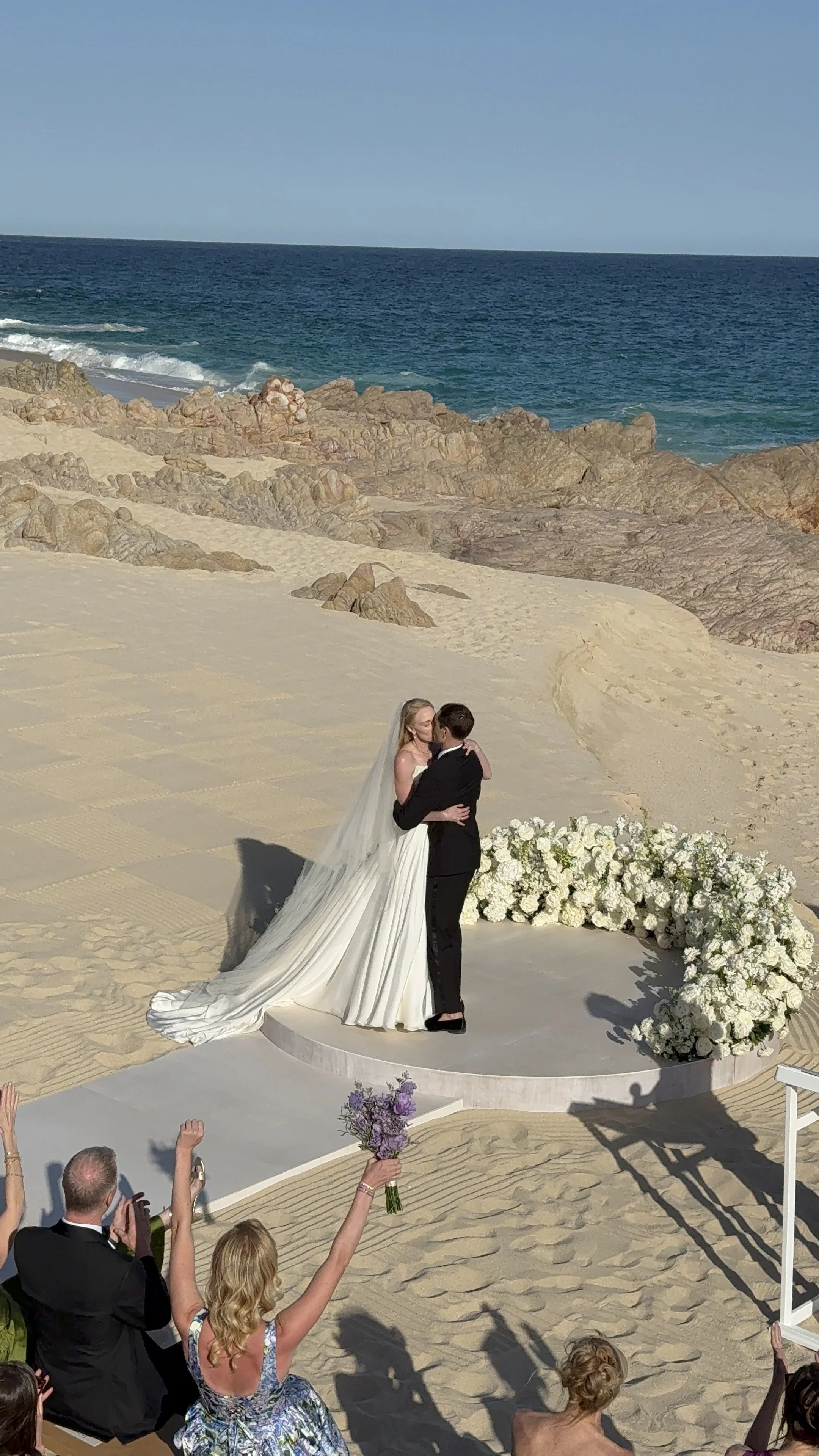 A bride and groom hugging during their beach wedding ceremony, with guests in the foreground celebrating. The ceremony platform decorated with white flowers is on sandy terrain by the ocean.