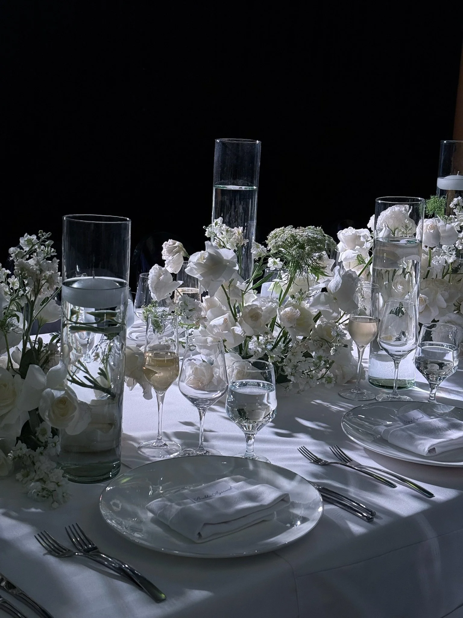 Elegant table setting with glassware, white flowers, and white plates on a white tablecloth, against a dark background.