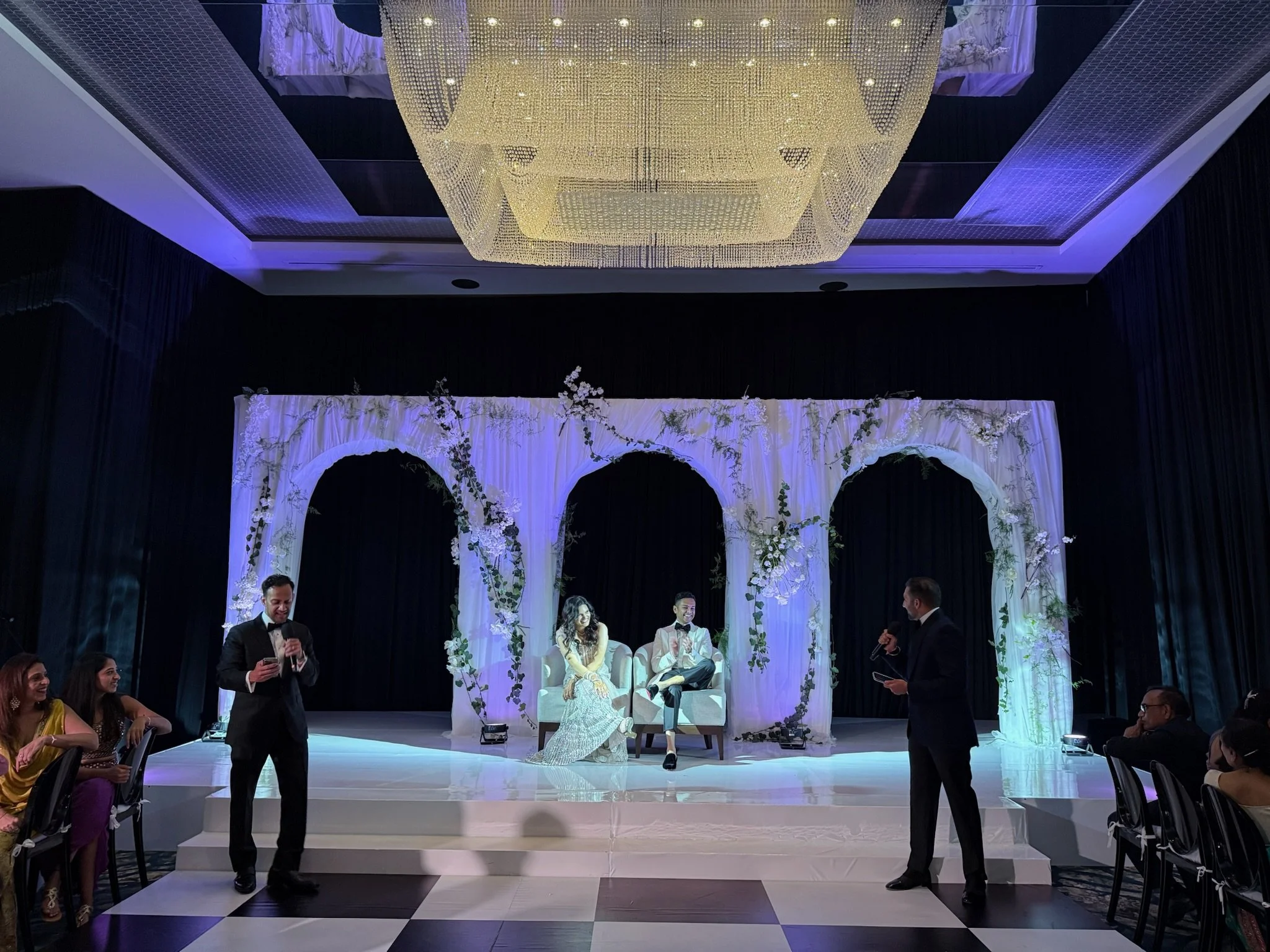 A wedding reception with a decorated stage featuring white flowers and drapery, where the bride and groom are seated. Two men in suits are standing and speaking into microphones, while guests sit at round tables watching. The ceiling has a large chan