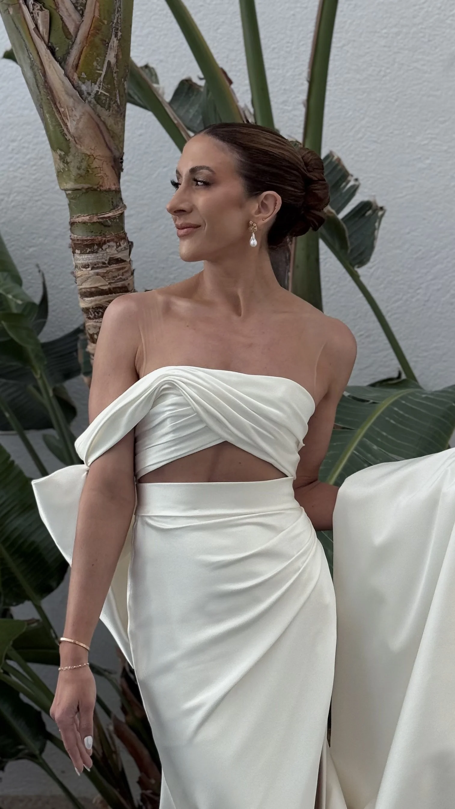 A woman in an elegant white dress with a cut-out midsection and off-shoulder design, standing outdoors near tropical plants, with her hair styled in a sleek bun and wearing earrings and a bracelet.