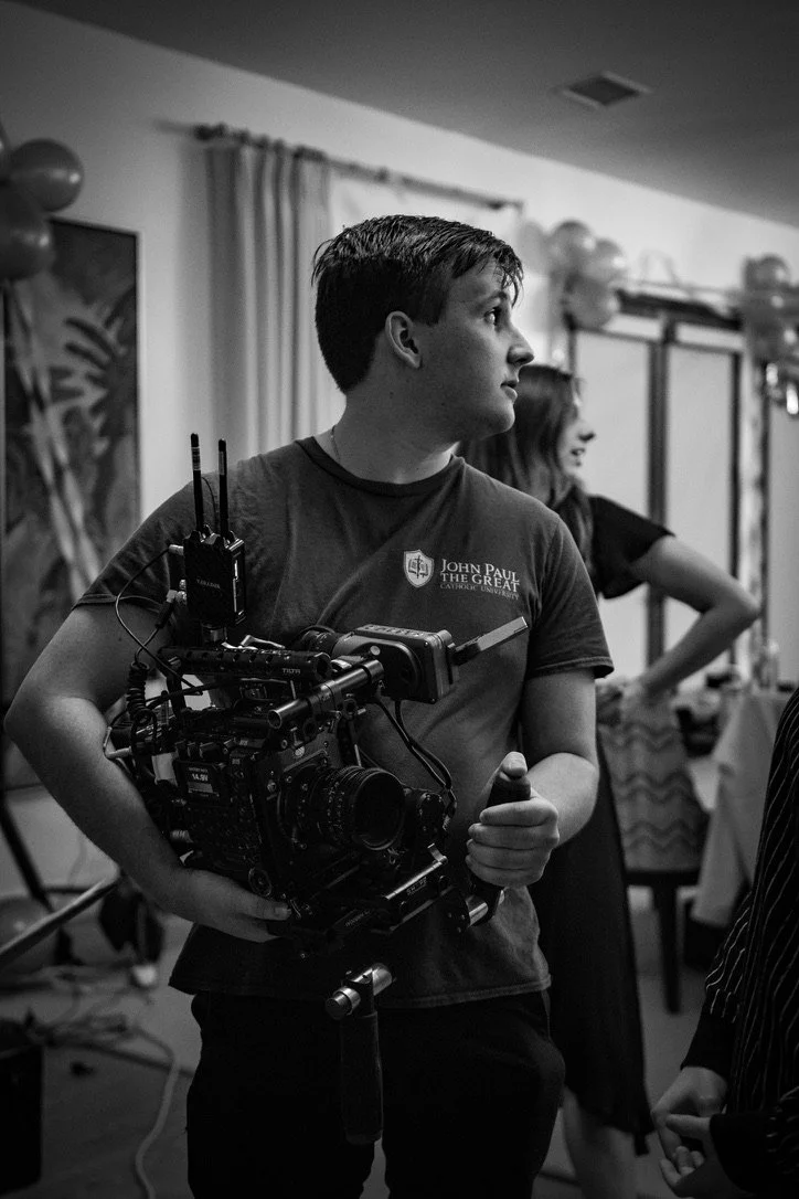 A young man holding a professional video camera, standing indoors at an event with balloons and curtains in the background.
