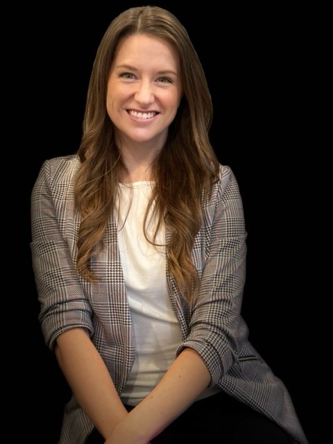 A smiling young woman with long brown hair, wearing a plaid blazer and a white shirt, sitting against a black background.