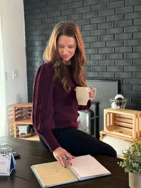 Woman with brown hair sitting at a table, holding a mug, looking at an open notebook.