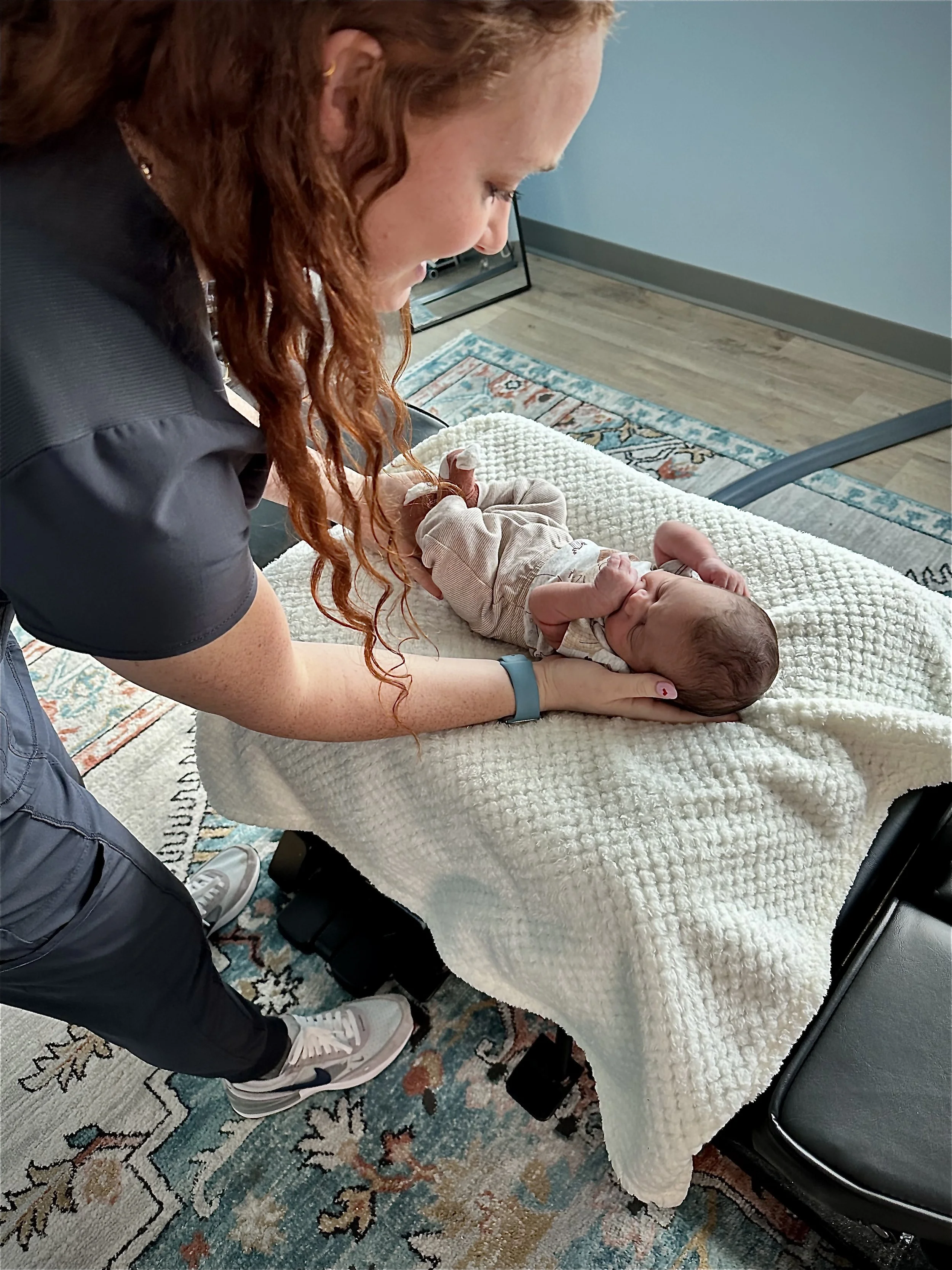 A woman with long red hair brushing her hair back while leaning over a newborn baby lying on a soft white blanket on a medical examination table. The woman is smiling and working on the baby using gentle specific movements.