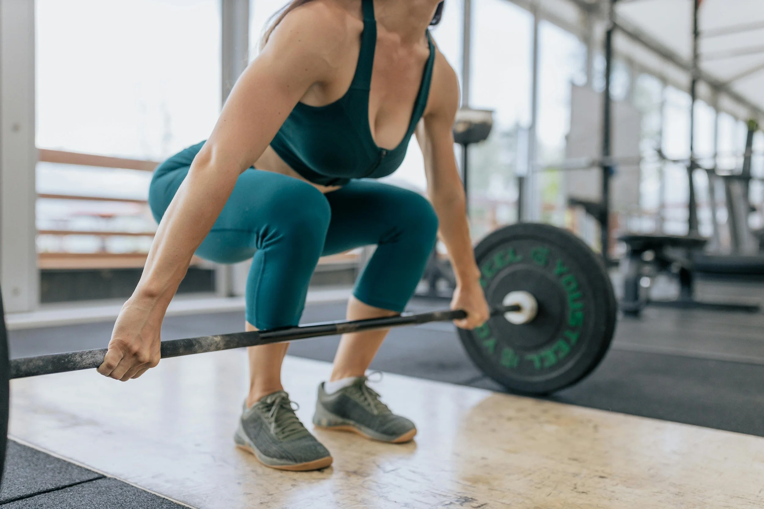 A woman in teal workout clothes performing a deadlift with a barbell inside a gym.