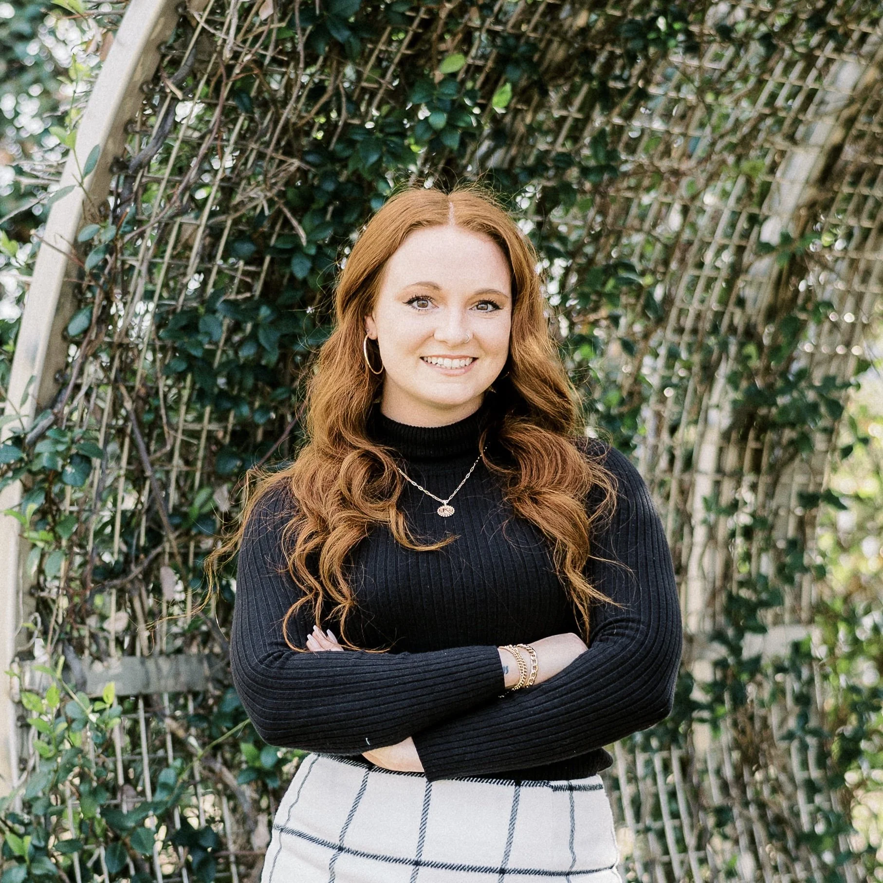 A woman with long red hair, wearing a black turtleneck and plaid skirt, standing with arms crossed in front of a garden arch covered in greenery.