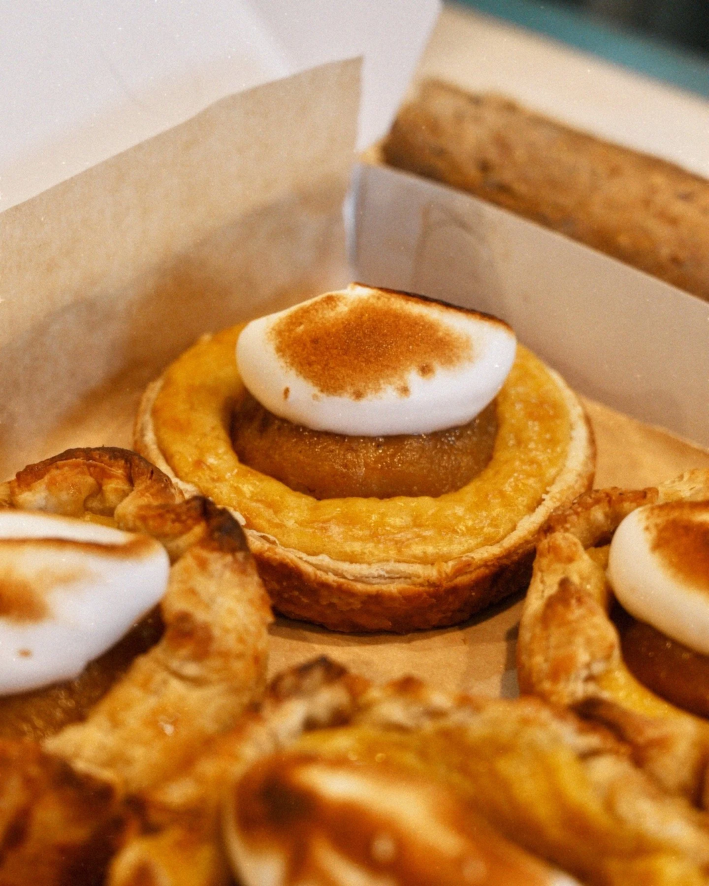 Close-up of a pastry with meringue, pastry crust, and a small container of lemon curd, with a box of cookies in the background