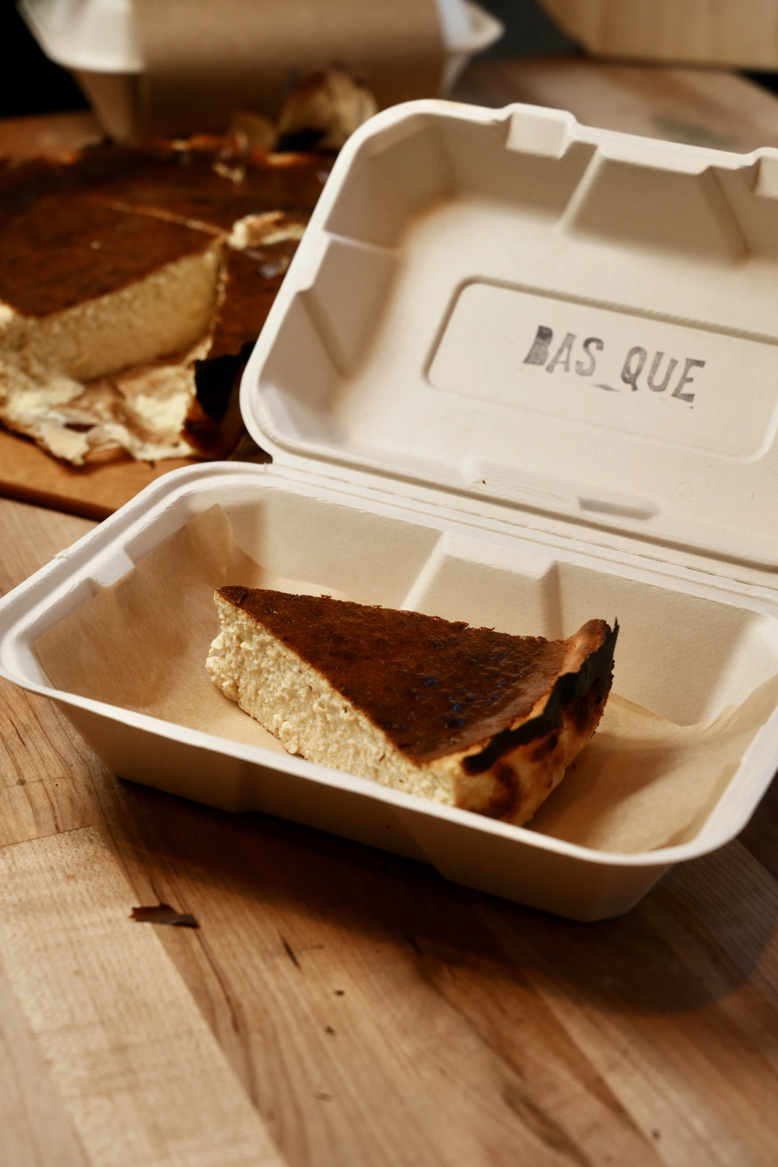 Slice of cheesecake in a white takeout container with a wood table surface in the foreground and a whole cheesecake in the background.