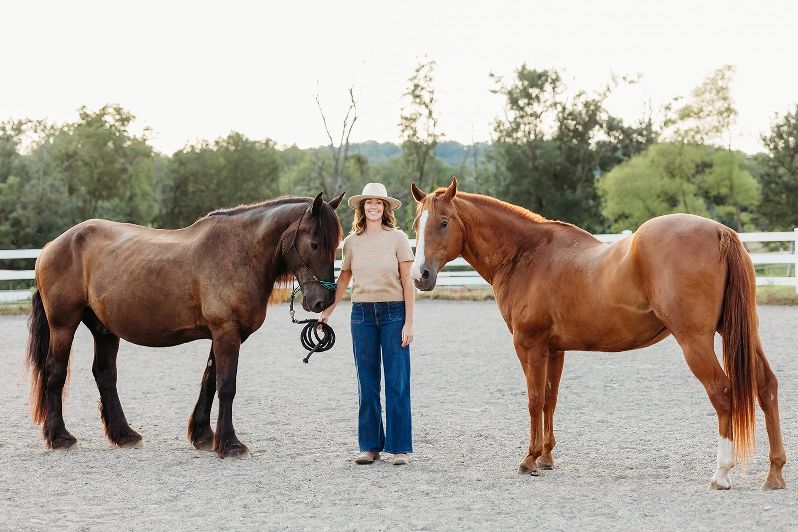 A woman wearing a beige sweater, jeans, and a white hat standing between two horses on a sandy outdoor riding arena, with trees and a white fence in the background.