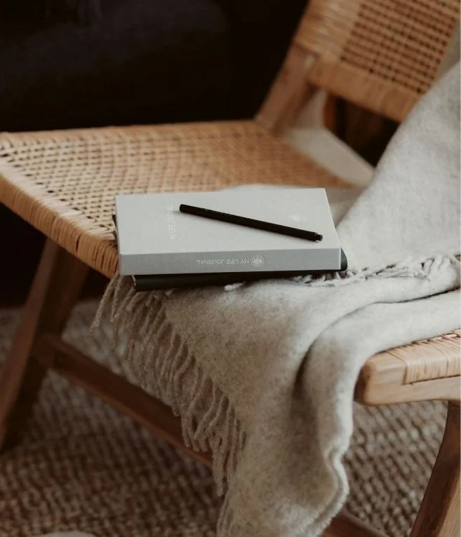 A black pen resting on a closed notebook placed on a cozy, fringed blanket on a wooden chair.