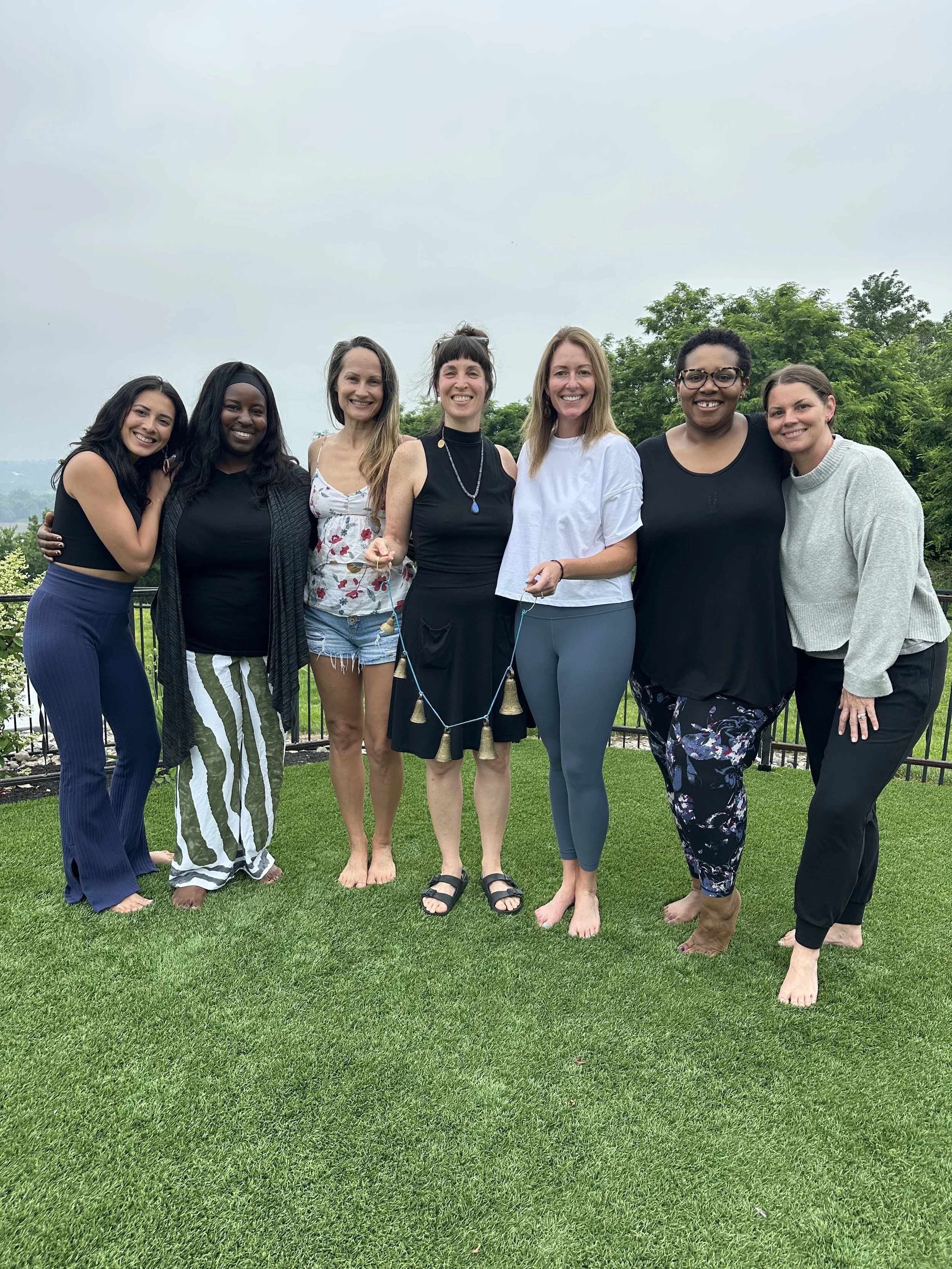 Group of seven women standing on a grass lawn, smiling, with trees and a cloudy sky in the background.