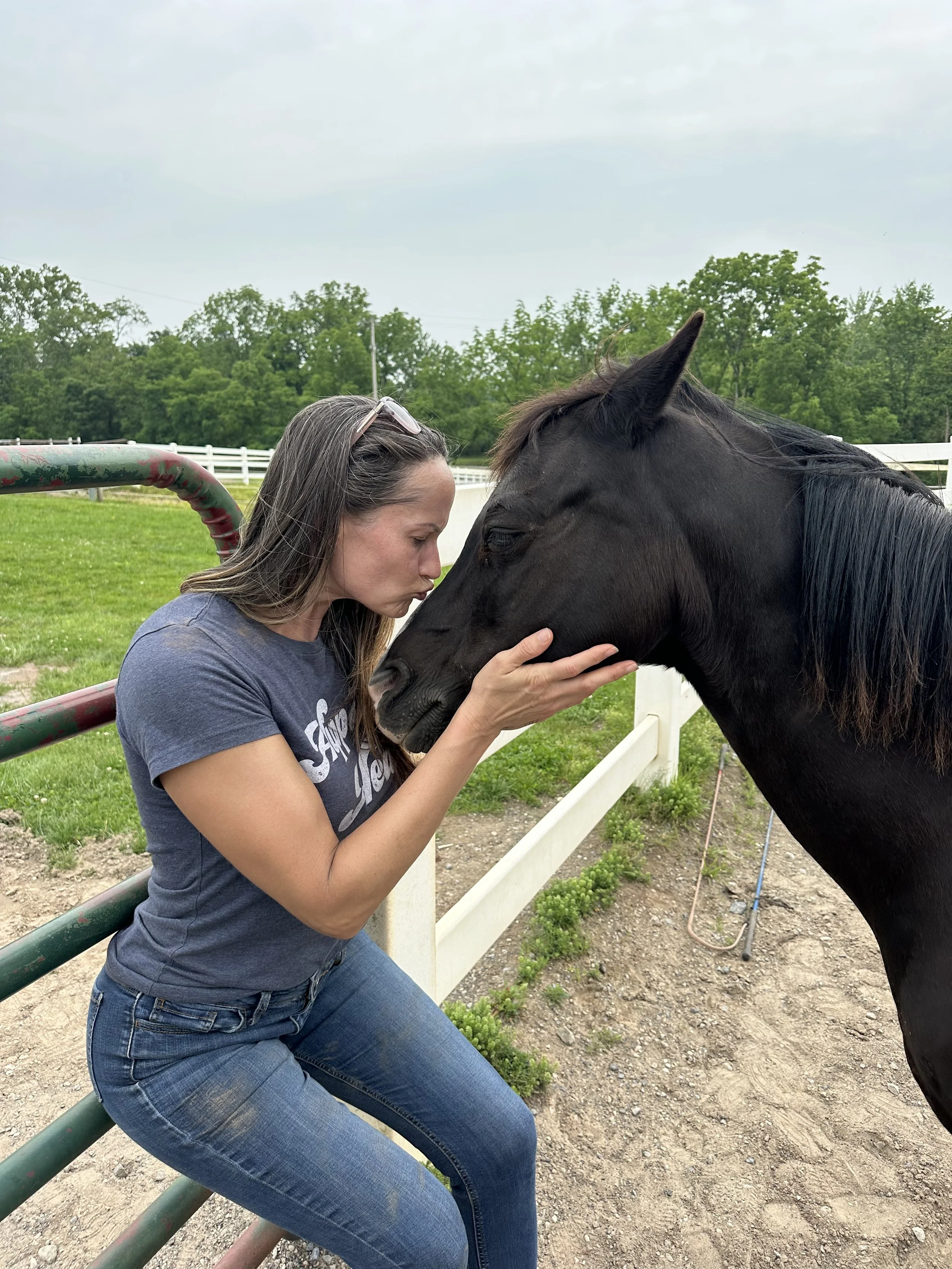 A woman with long hair, wearing a gray t-shirt and jeans, leans over a fence to gently hold and kiss a black horse's face at a farm with green grass and trees in the background.