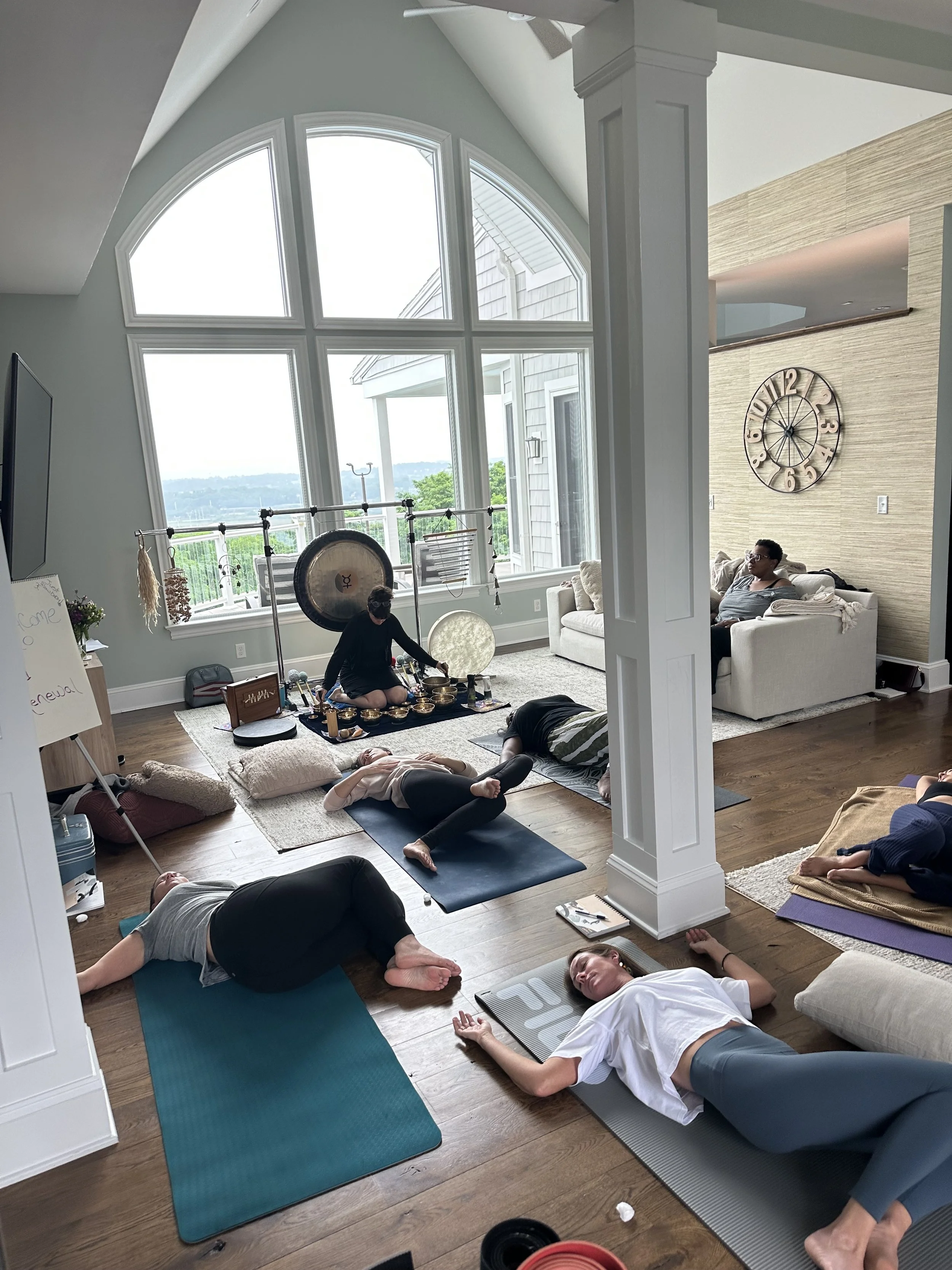 People participating in a yoga or meditation class in a living room with large windows, hardwood floors, and relaxing decor.